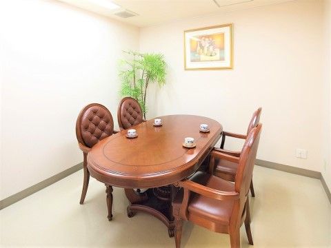 白い壁に囲まれた部屋には、高級感のある楕円形のテーブルと椅子がある。テーブルの上にはティーカップが置かれ、壁には絵画が飾られている。