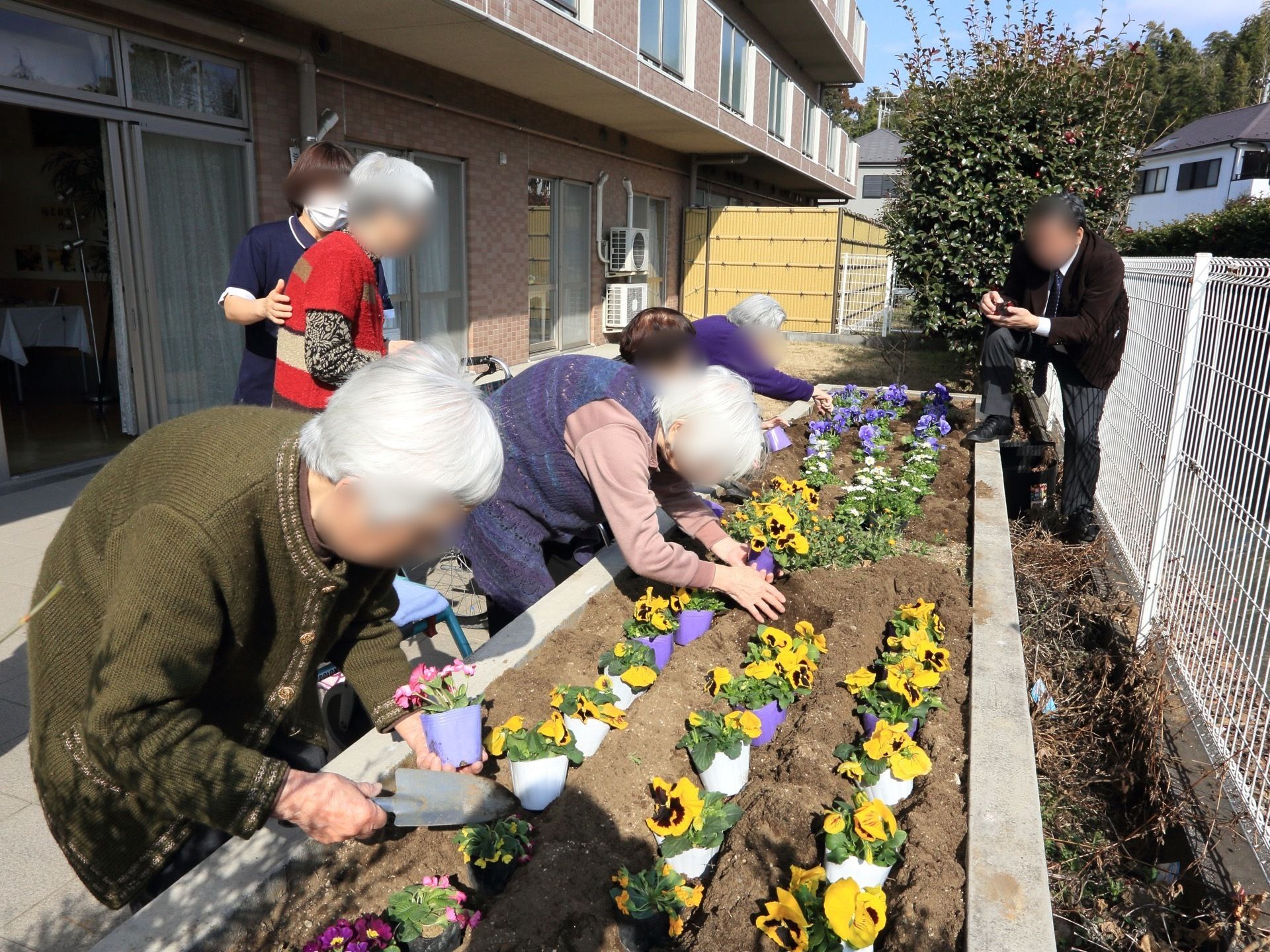 敷地内の花壇の様子。５人のご入居者がパンジーなどの花を植え替えている。また、スタッフが介助している姿が見える。