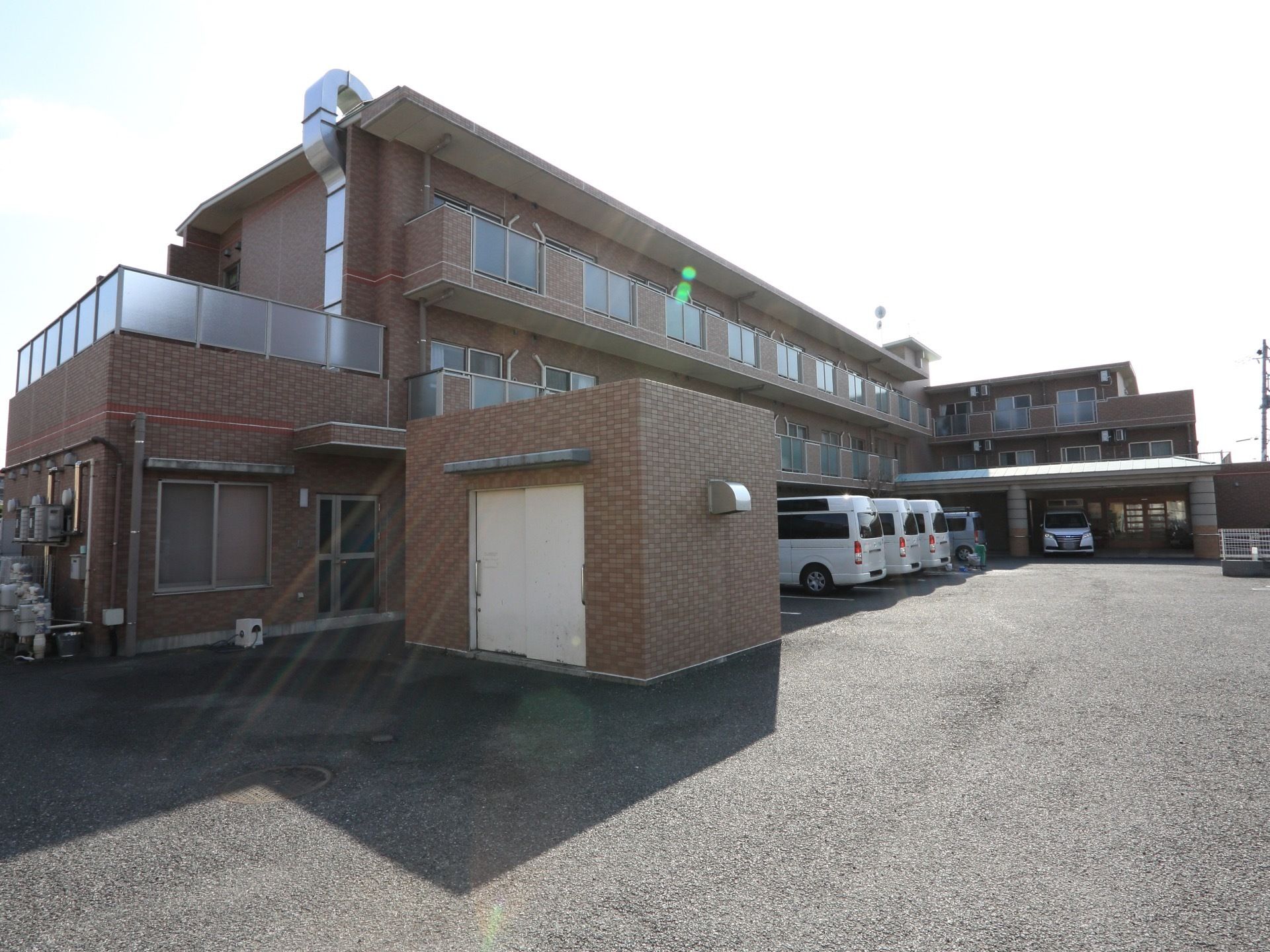 茶色のレンガ調の外装で３階建ての建物。施設の前に広い駐車場がある。２階と３階にはベランダがついている。