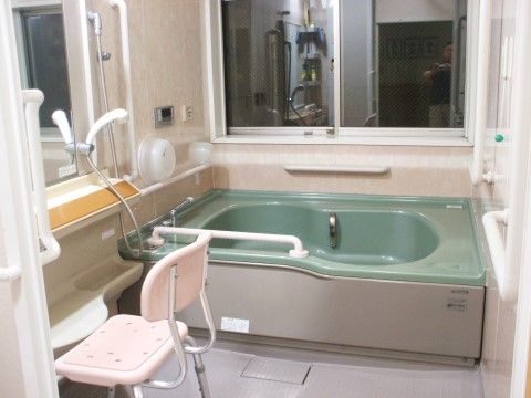 上部が濃い緑色の浴槽のある浴室。洗い場にはシャワーチェアが置かれている。要所に手すりが設置されている。