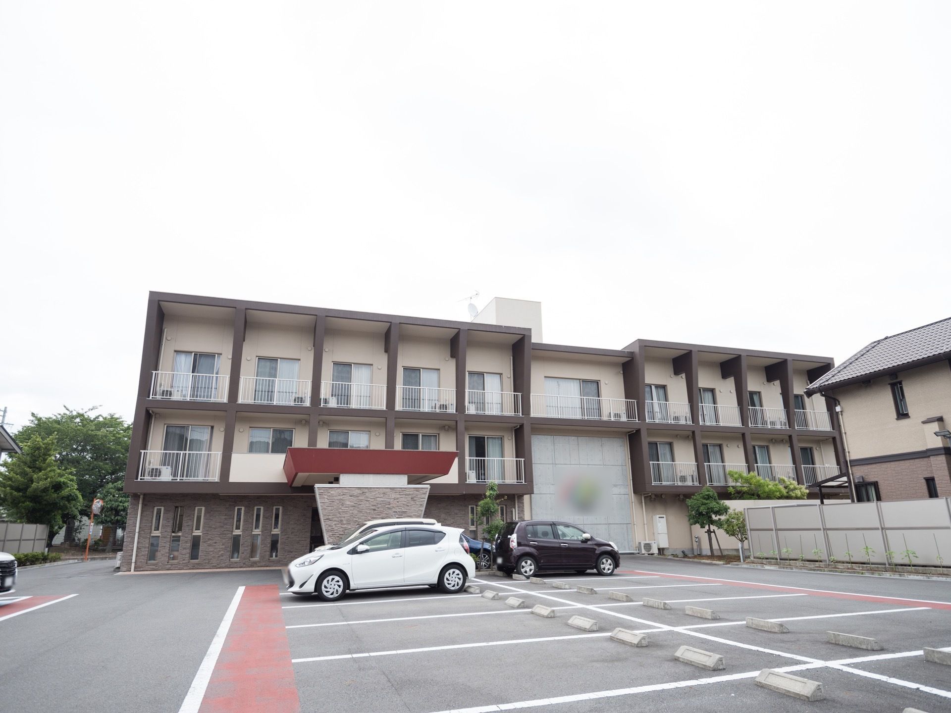広い駐車場の前に、3階建ての施設が建てられている。2階、3階には、それぞれベランダがあり、柱で区切られている。