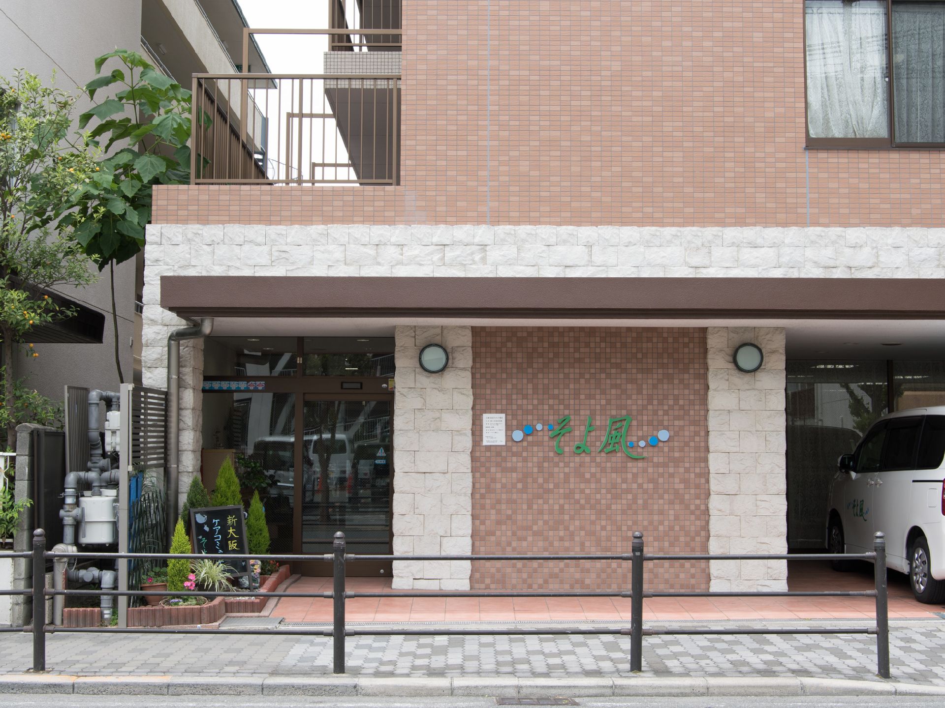 施設の出入り口は自動ドアになっていて植木が植えられている。レンガ調の壁には緑で施設名が大きく記載されている。横には駐車場もある。