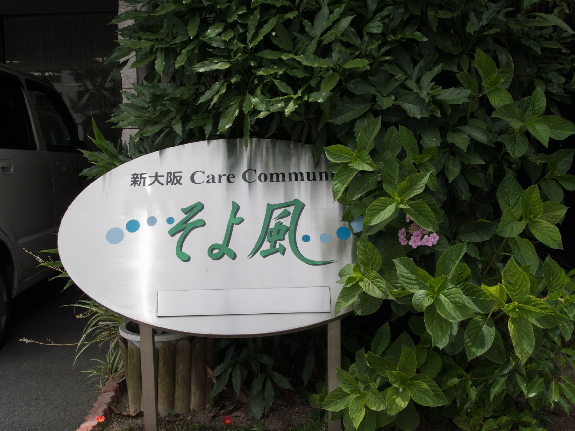 施設の駐車場の脇には植木が植えられている。植木の中には銀色の金属パネルに青と緑で施設名とロゴが描かれた看板が立てられている。