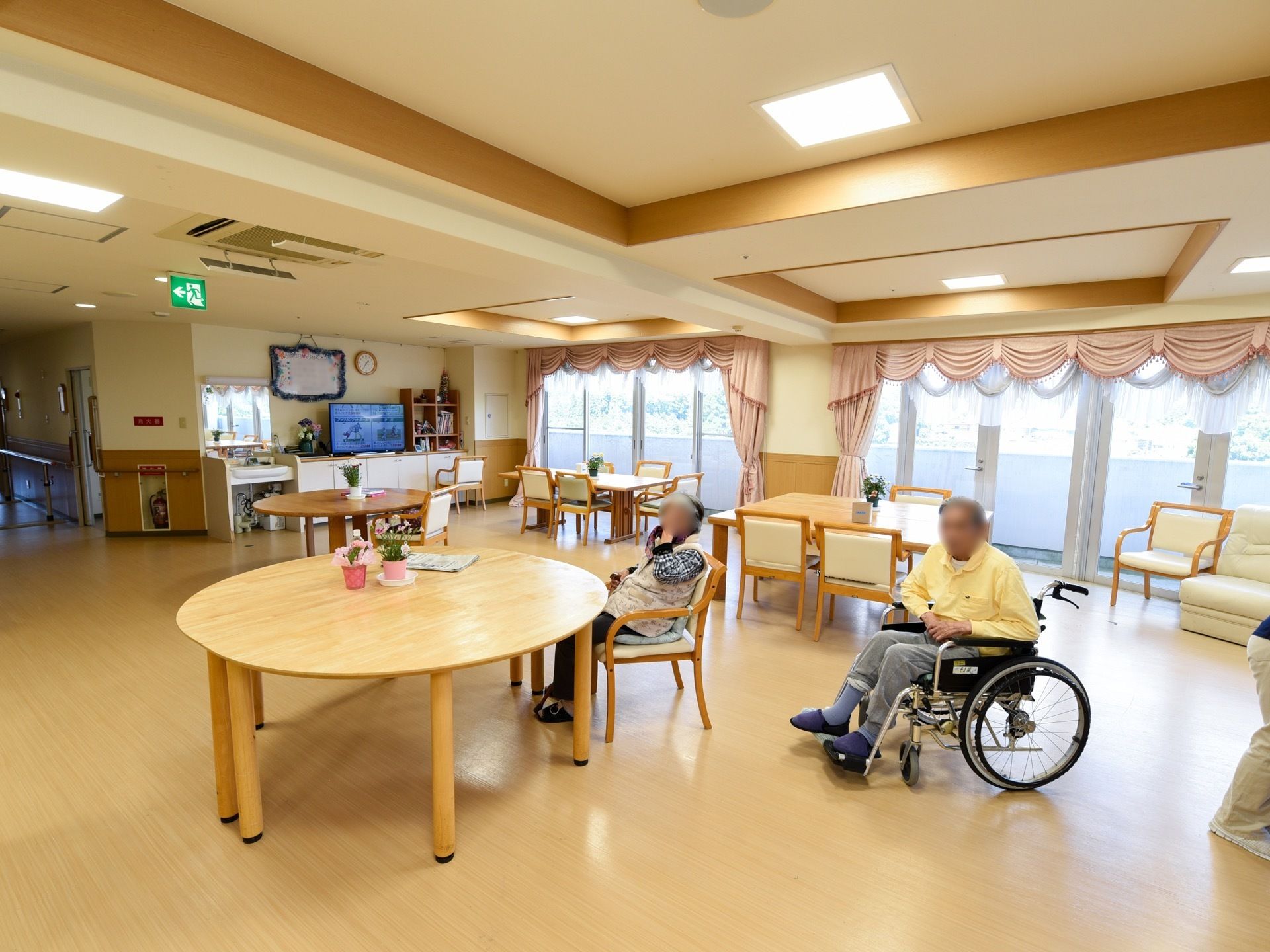 フローリングの敷かれた明るい室内で、男女のご入居者が椅子に座ってくつろいでいる。室内の各テーブルには小振りの観葉植物が飾られている。