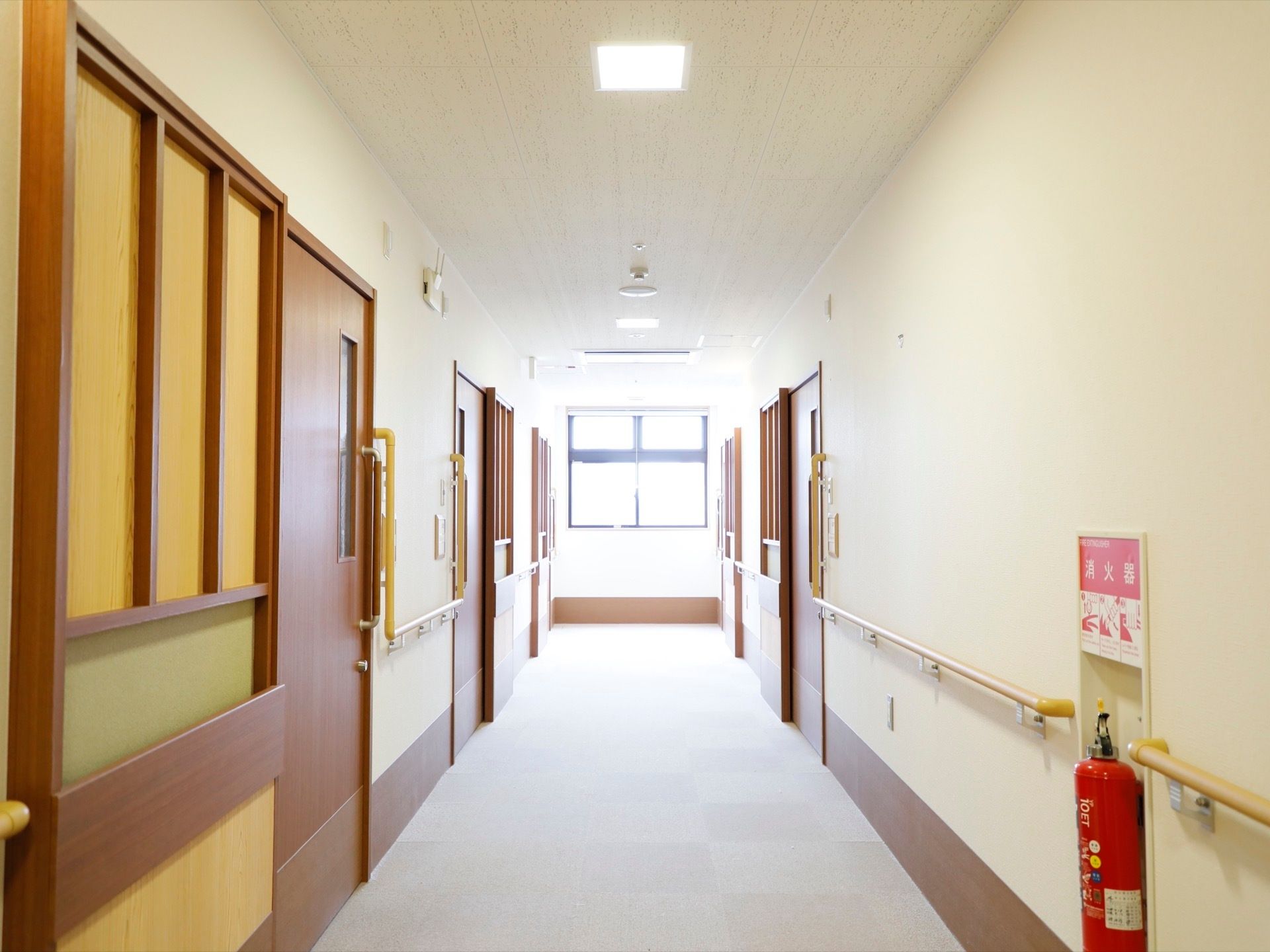 通路正面には大きな窓があり、陽射しが差し込んでいる。居室のドアが５枚見えている。消火器が設置してある。