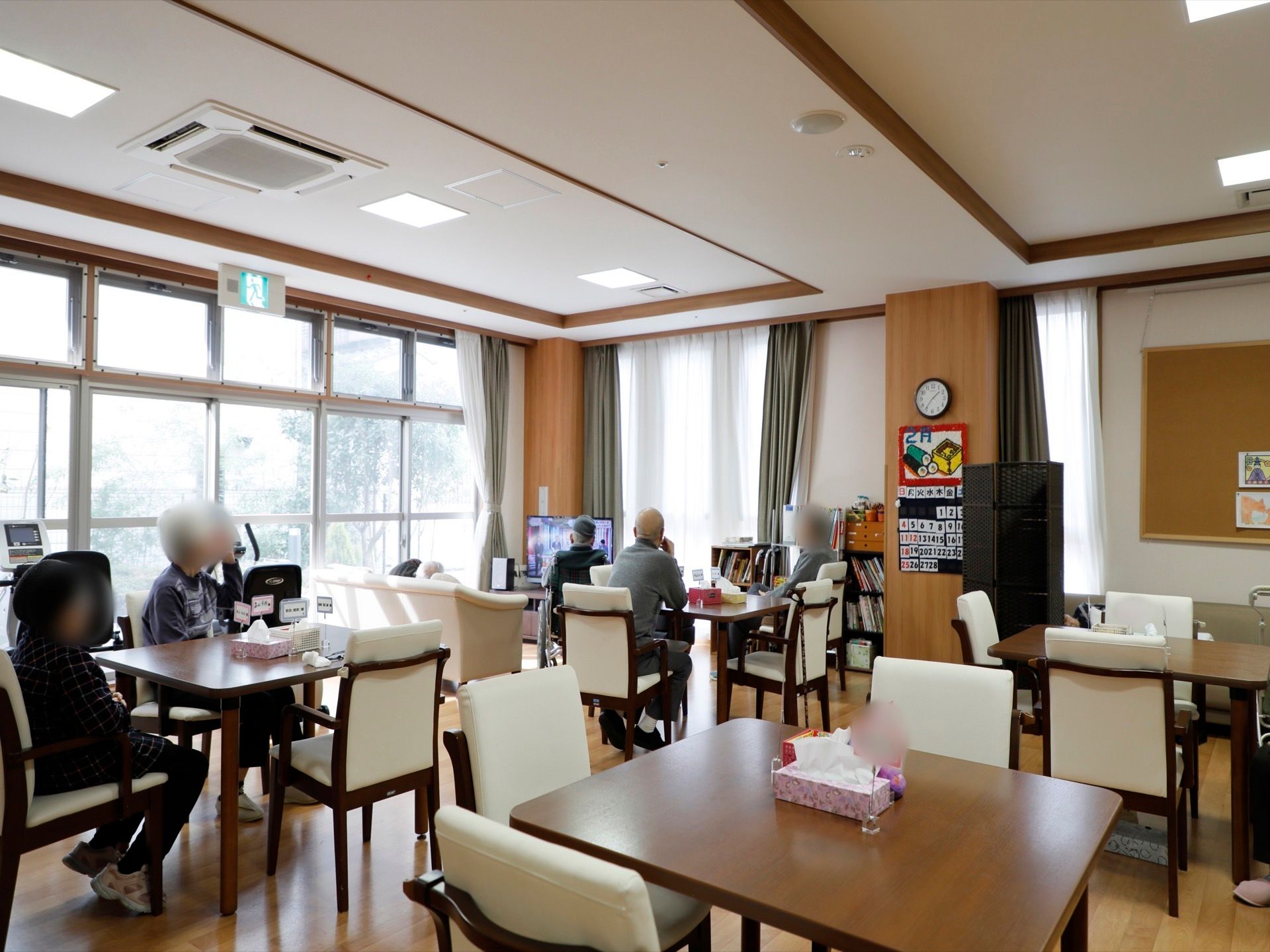 ダイニングルームには、正方形のテーブルと椅子が配置されている。入居者様は好きな場所に腰かけ、備え付けのテレビを見てくつろいでいる。