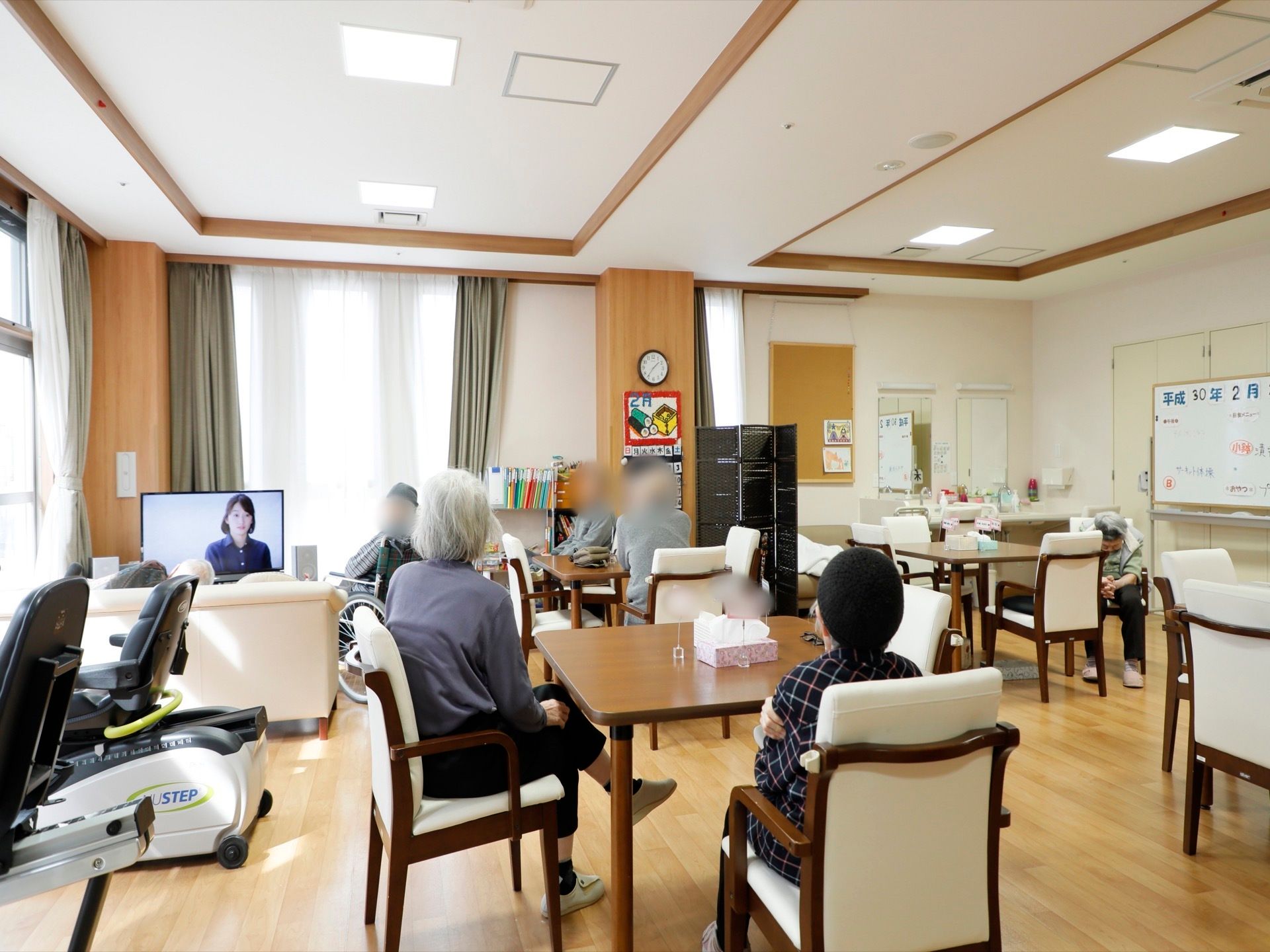 共有スペースの様子。木目調の内装で、落ち着きのある雰囲気が漂う。テレビやソファが置かれており、入居者様がくつろいで過ごしている。