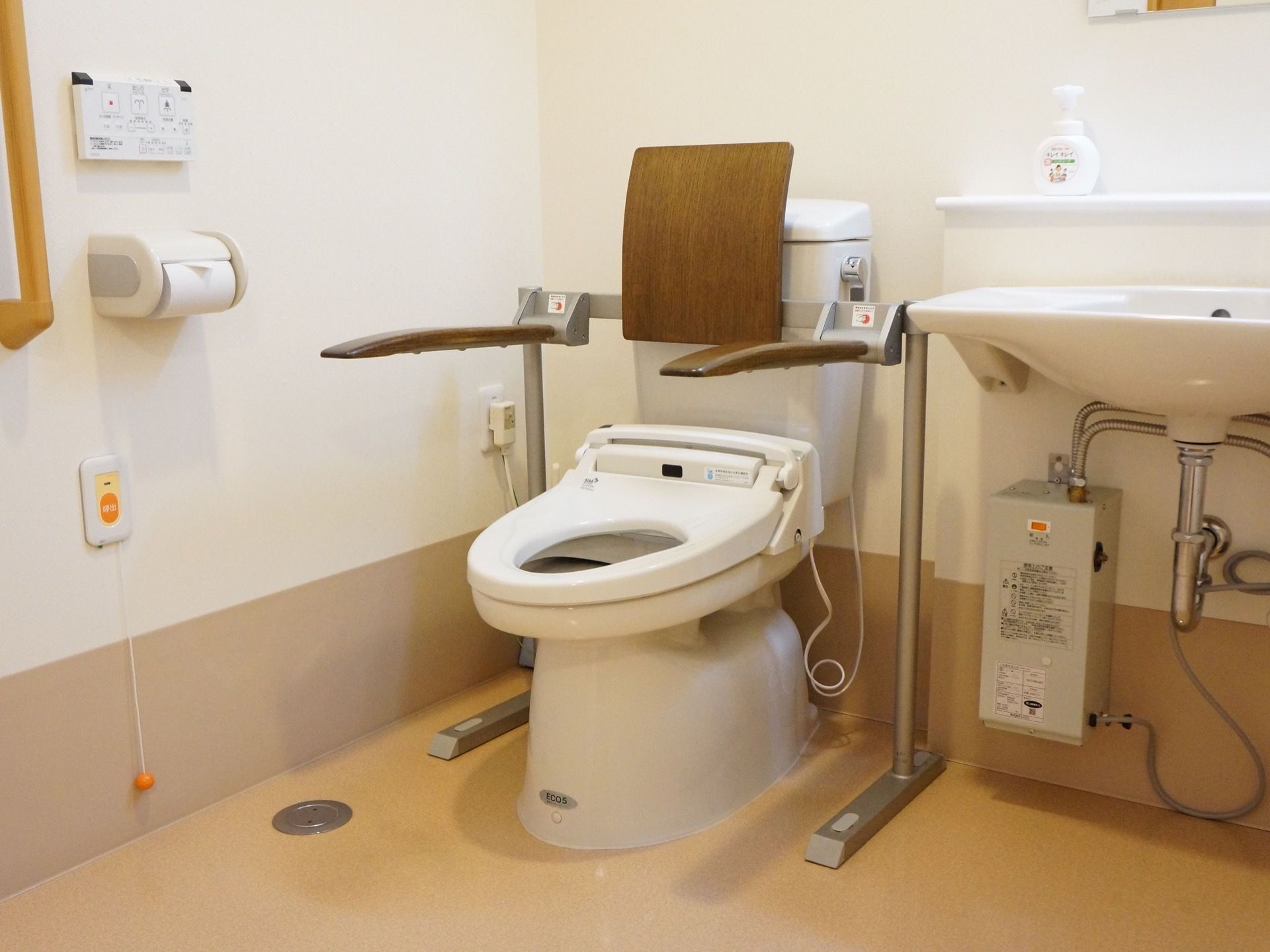 洗面台が備わっているトイレである。便器には、背もたれ付き肘掛け手すりが設置されている。呼び出しボタンが壁に取り付けられている。