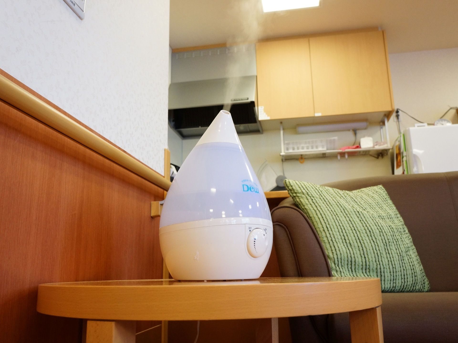 壁際ある丸いローテブルの上に加湿器が置かれている。奥にキッチンが設けられており、右横には茶色いソファがある。