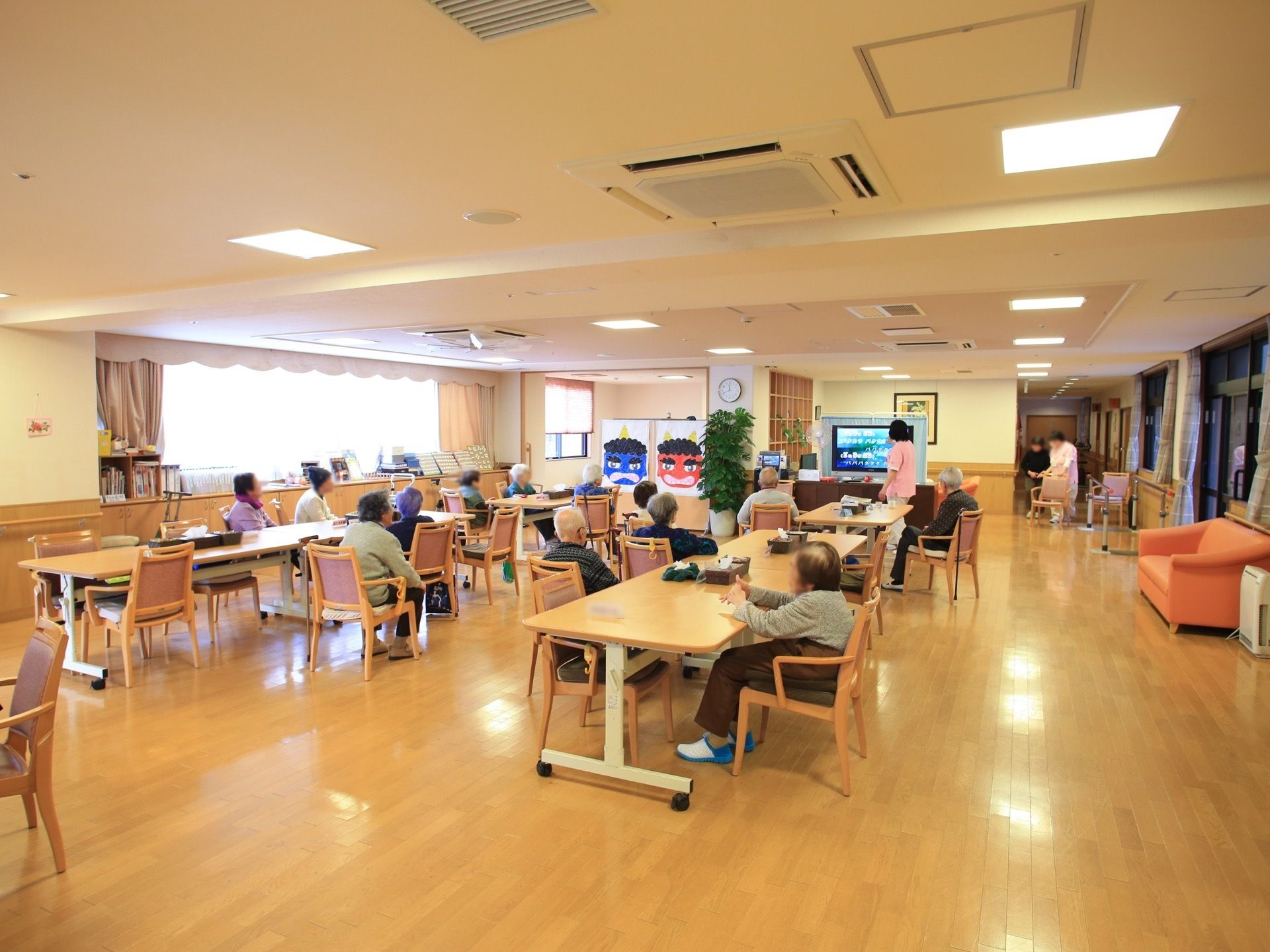 広々とした空間に、テーブルと椅子が並べられている。入居者様が集まり、レクリエーションが行われている。季節の行事のイラストが飾られている。