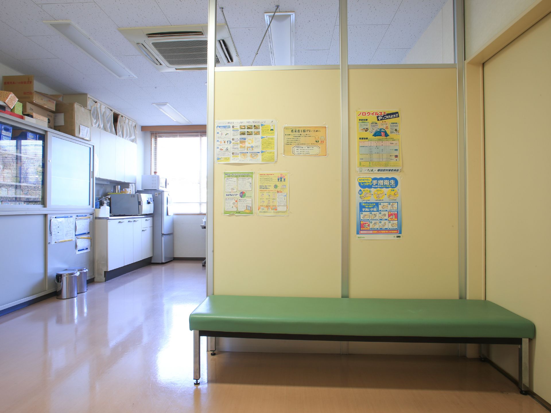 緑色の病院にあるようなベンチが置かれている。流し台とキャビネットがあり、壁には衛生に関するお知らせが貼られている。
