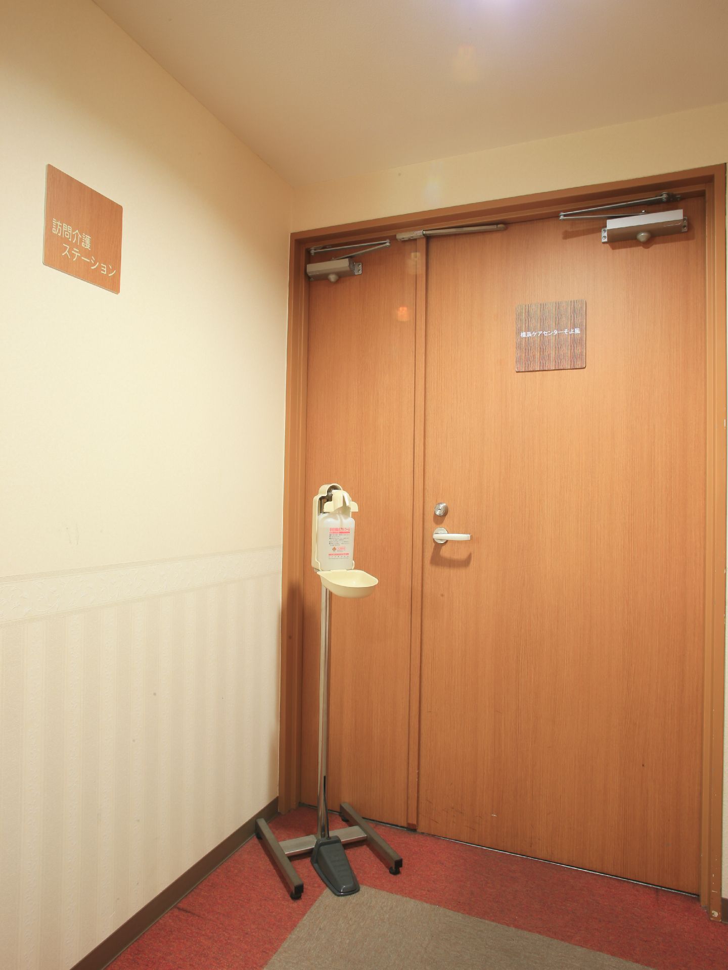 訪問介護ステーションの部屋へと続く、木目調のドアがある。側には消毒液がセットされたスタンドが用意されている。