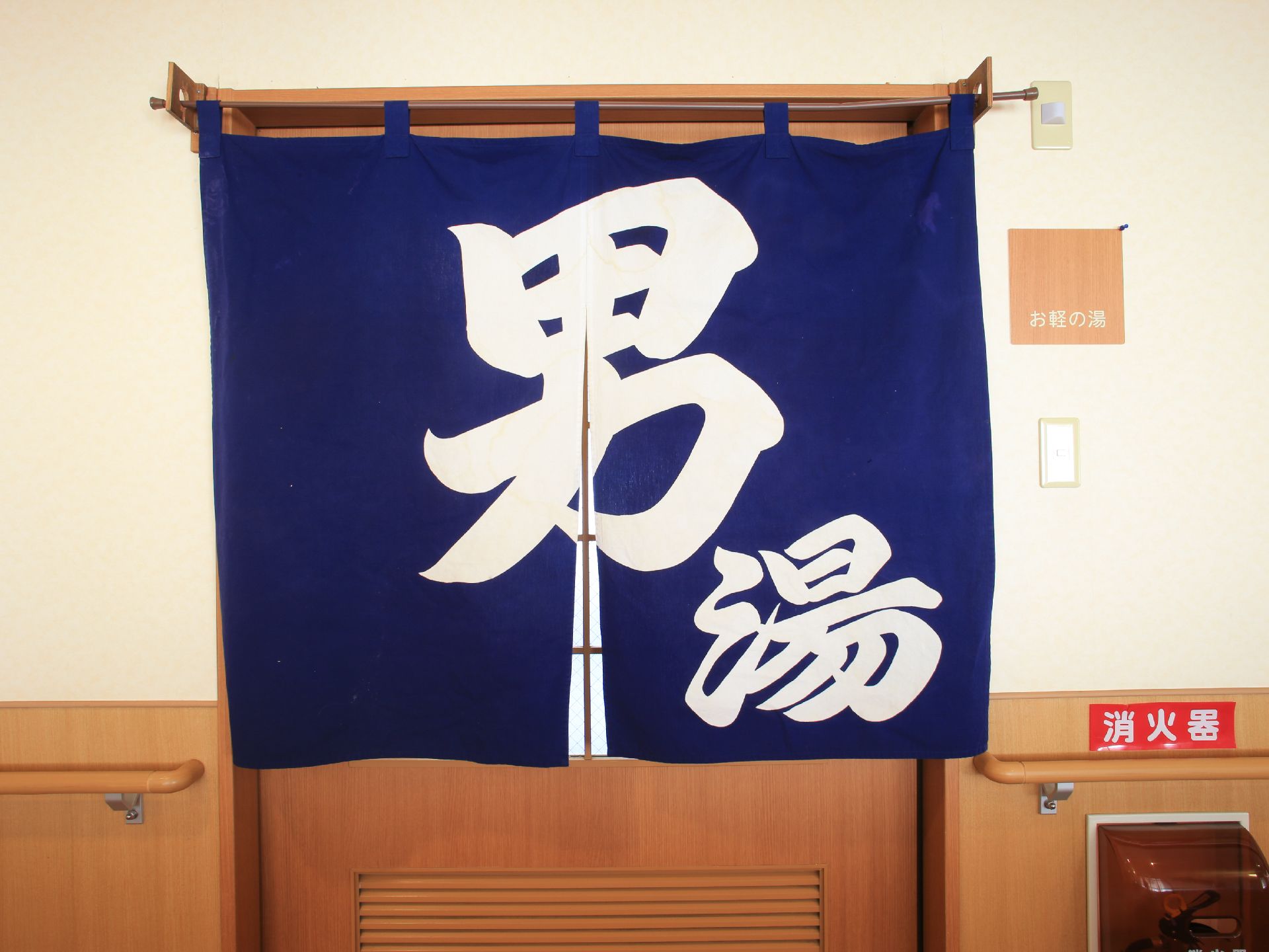 男湯と書かれた青い暖簾がドアにかかっている。幅のある横開きのドアである。入口の横には消火器が備えつけられている。