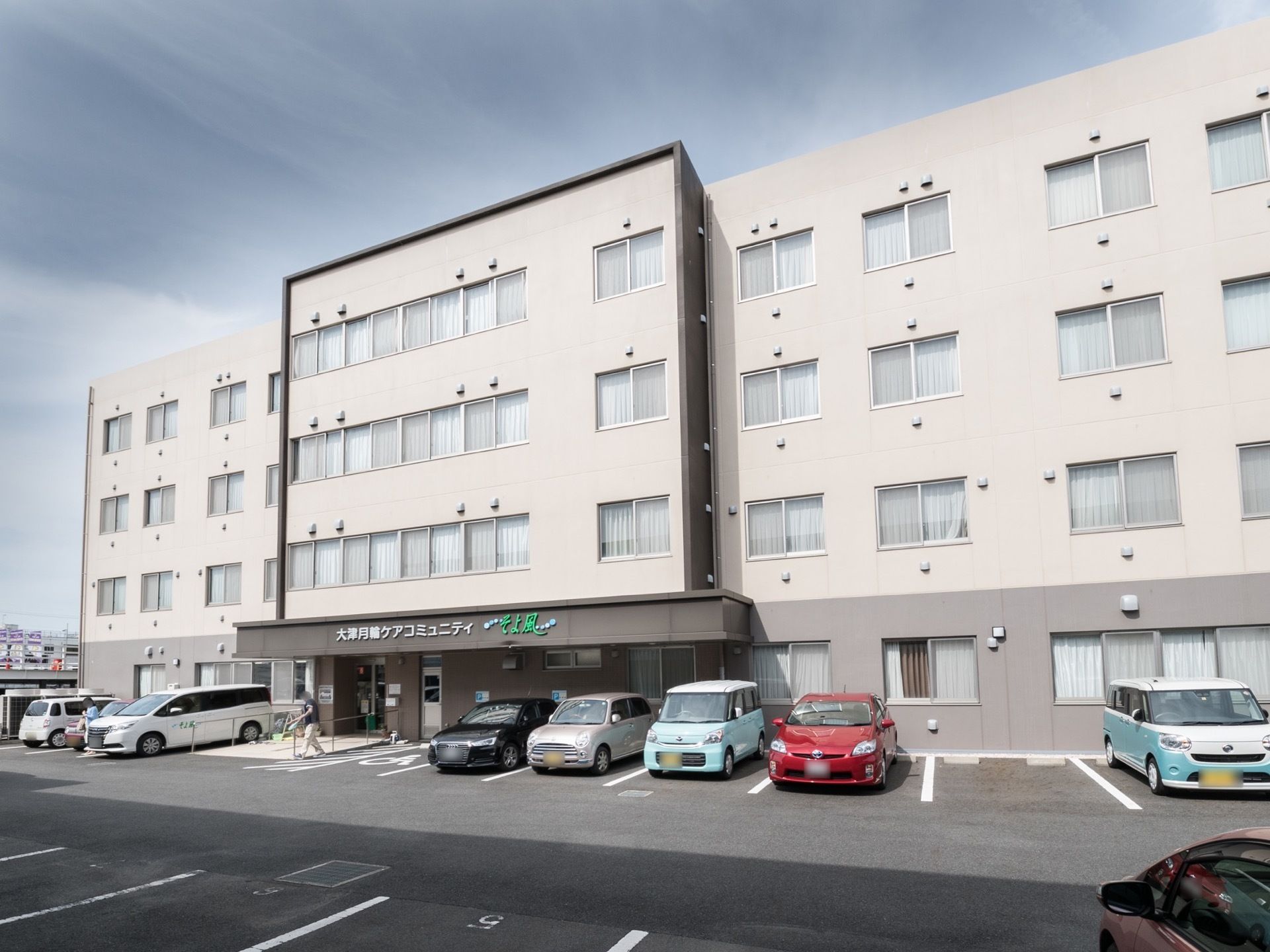 ベージュとグレーのモダンな４階建ての建物である。施設の前には広い駐車スペースがあり、車が多く駐車している。