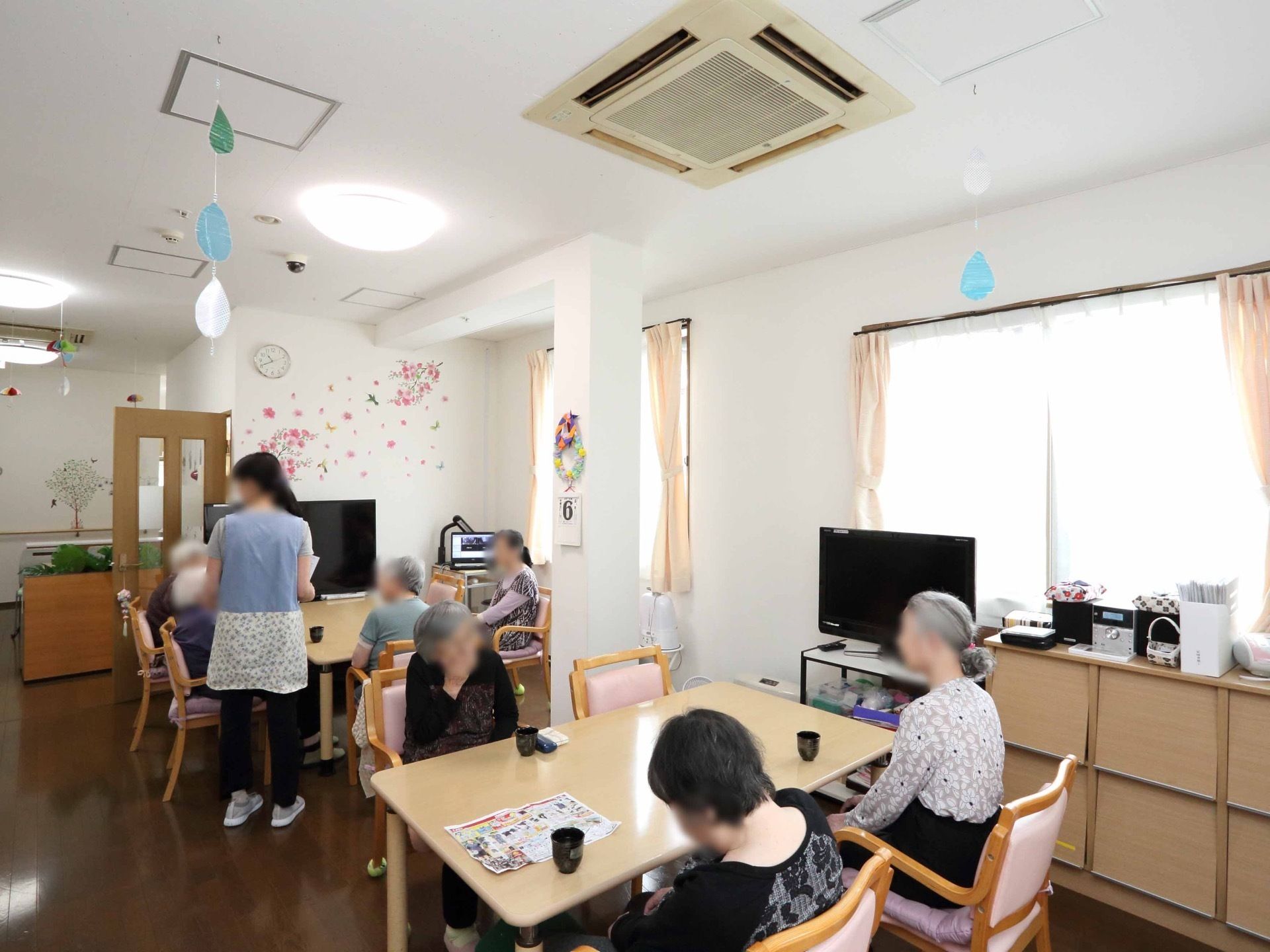 リビングスペースにはテレビが２台あり、各テレビの前にはテーブルと椅子が配置されている。着席している入居者様の前には湯飲み茶わんが置いてある。