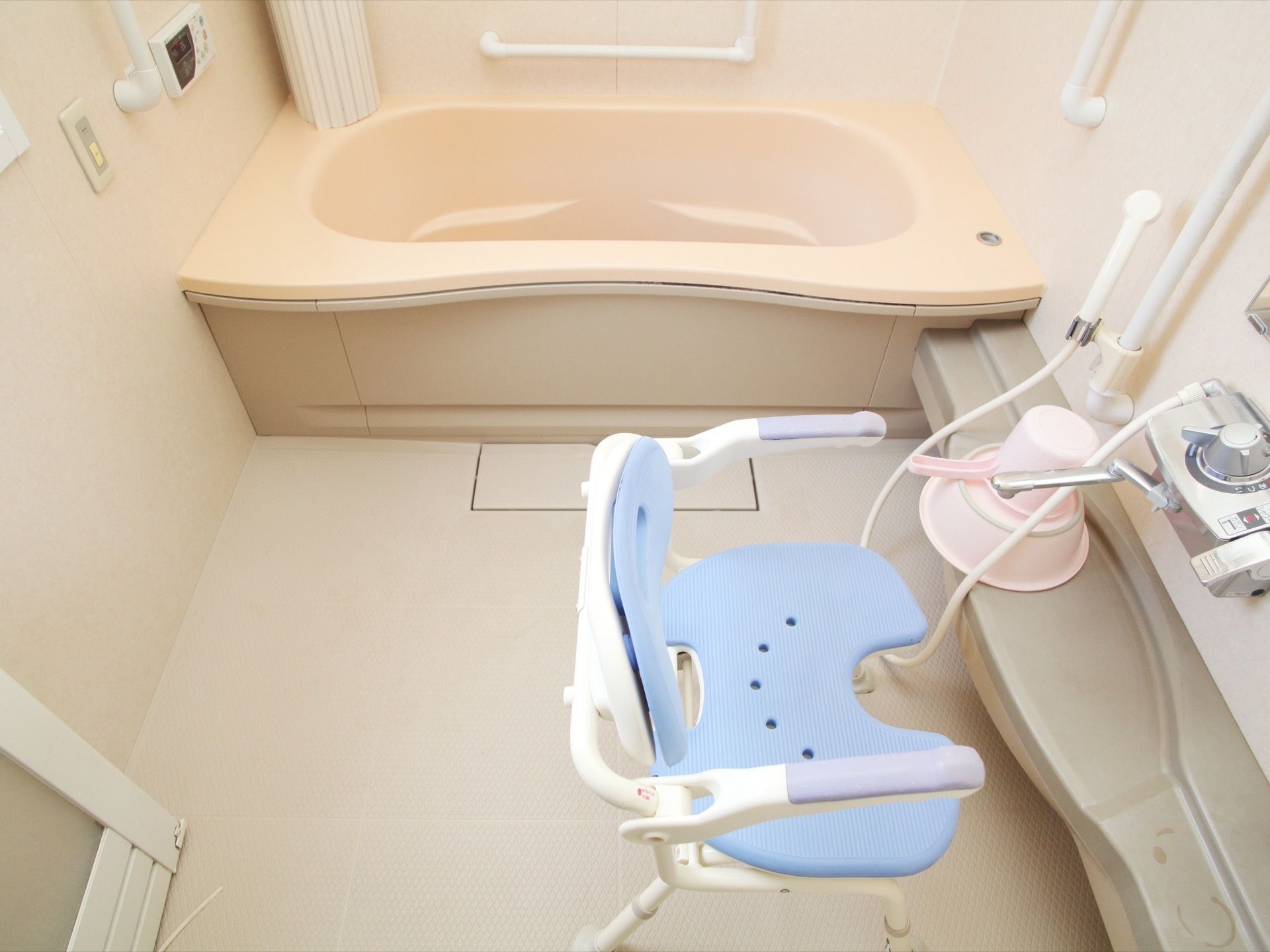 水色のシャワーチェアやピンクの洗面器、手桶が置かれている。ドアは中折れドアになっている。壁には操作パネルが設置されている。