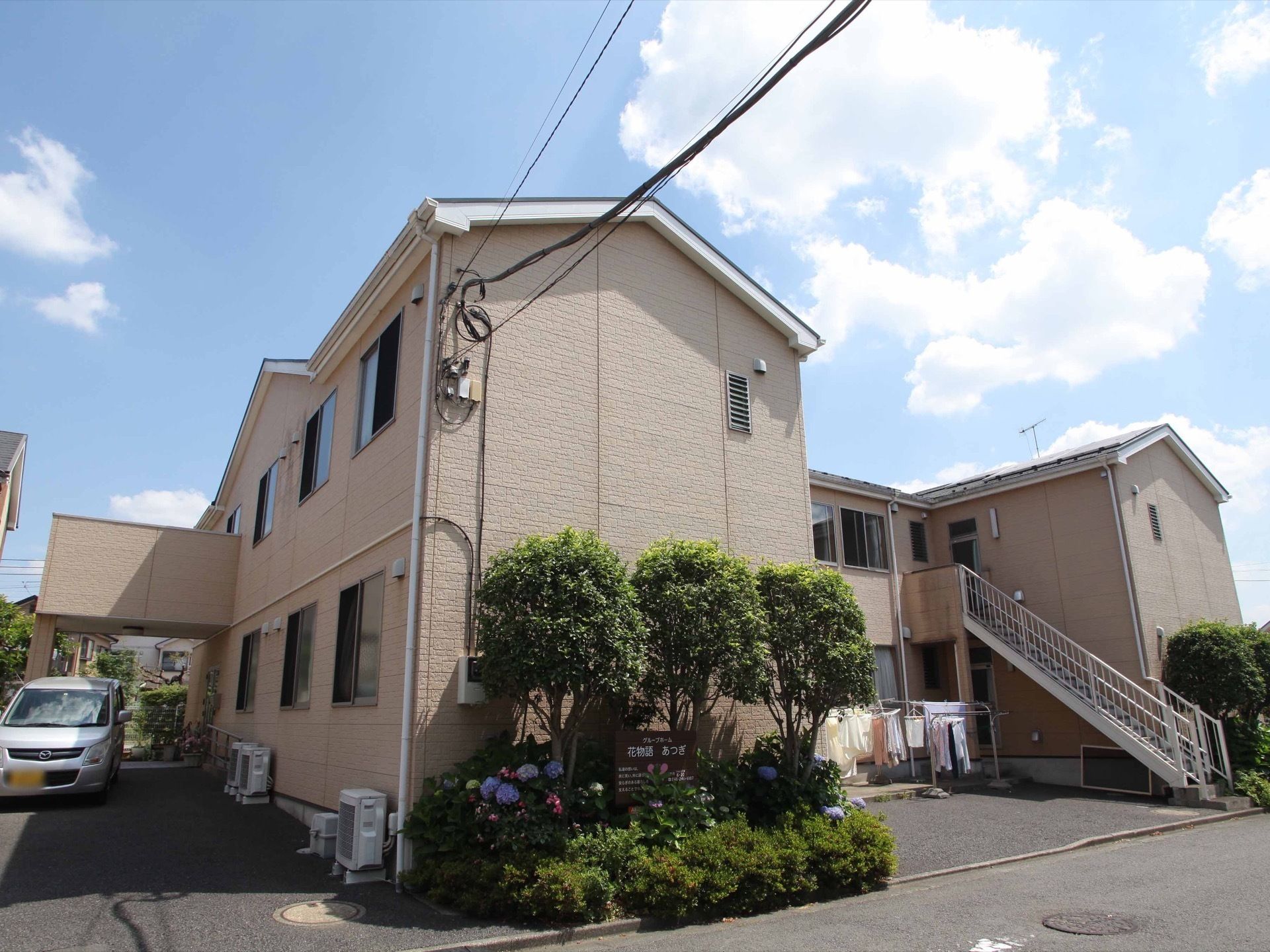 ２階建ての建物で、外階段がある。建物の前には花壇があり、木や花が植えられ、施設名の書かれた看板がある。