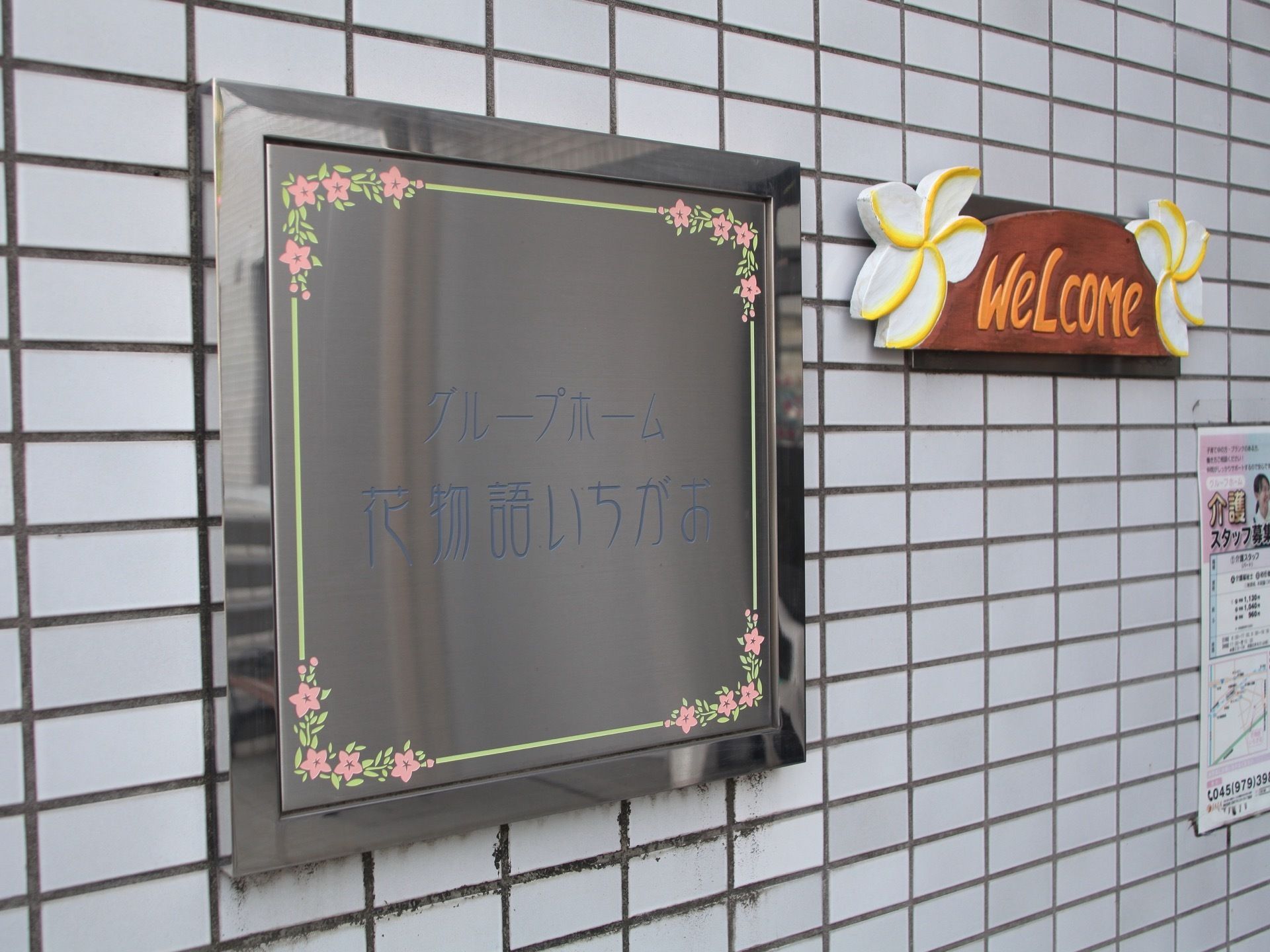 白色の長方形のタイルが施された外壁に、施設名や花の装飾が記されたシルバーのプレートが掲げられている。