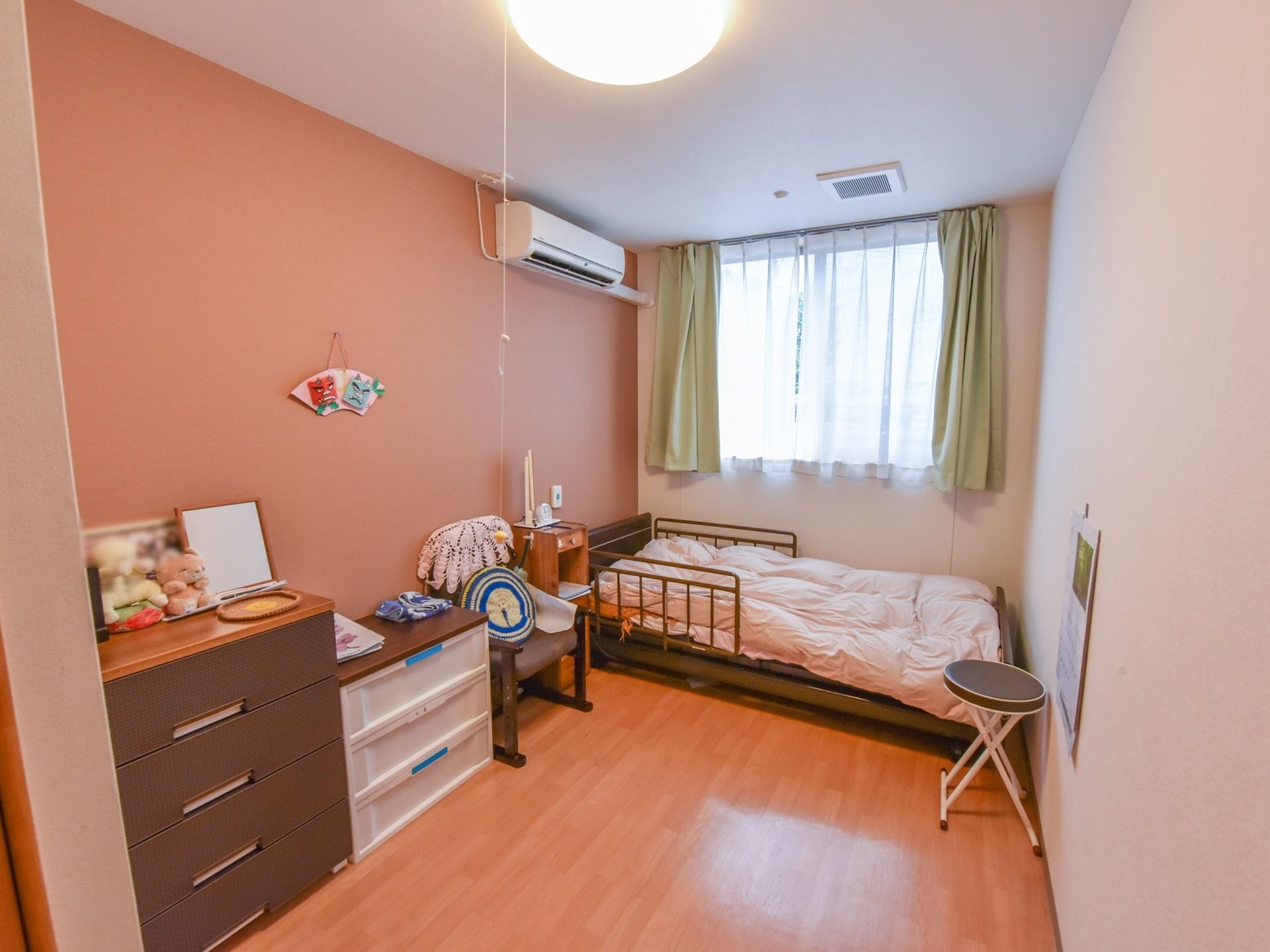 居室は白とブラウンを取り入れ、ぬくもり感が漂っている。窓際に介護用ベッドを配置し、壁際に収納家具や椅子などを置いている。