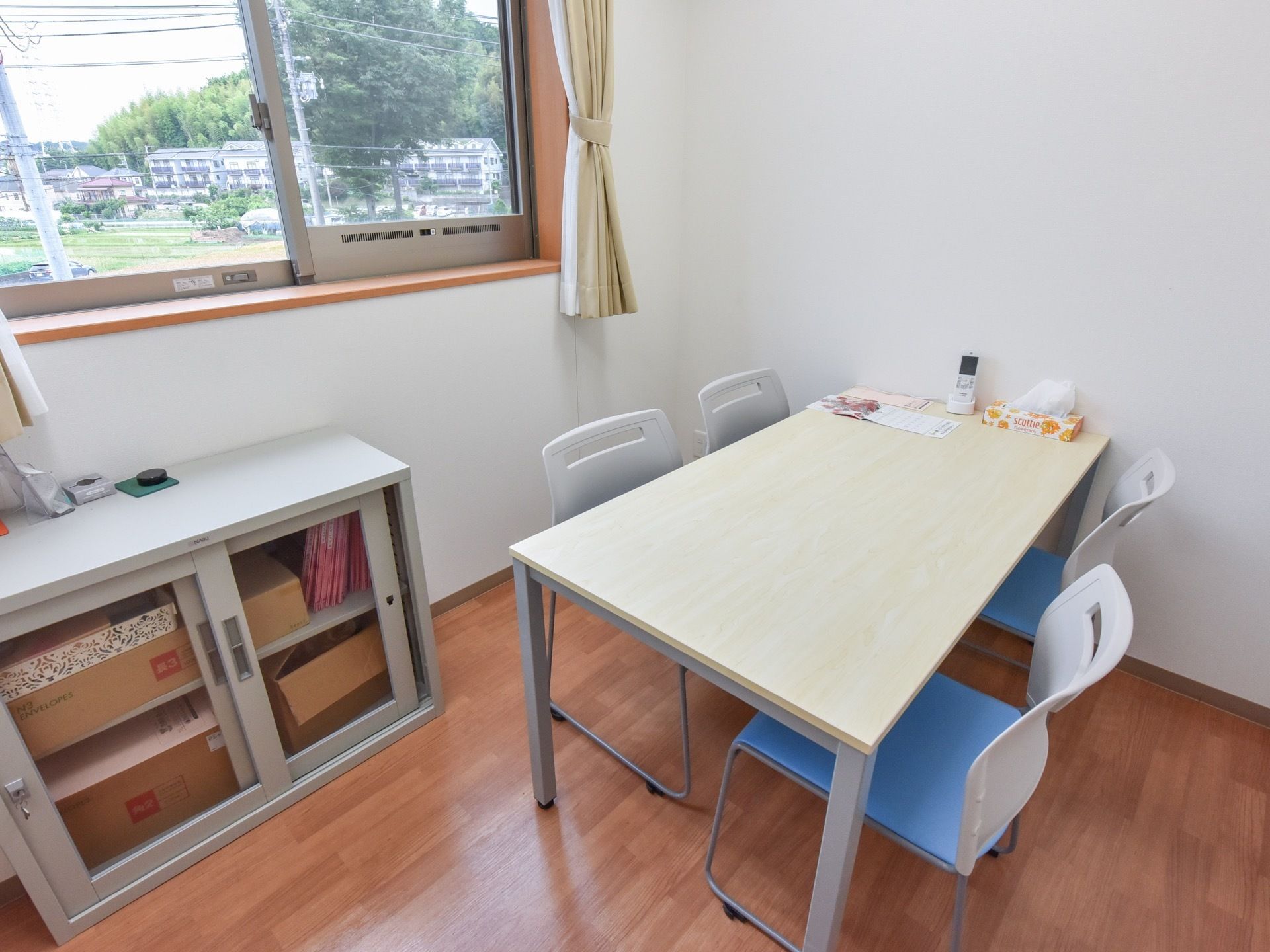 白い壁の部屋の窓からは建物や山が見えている。テーブルセットとファイルなどの入ったキャビネットがある。