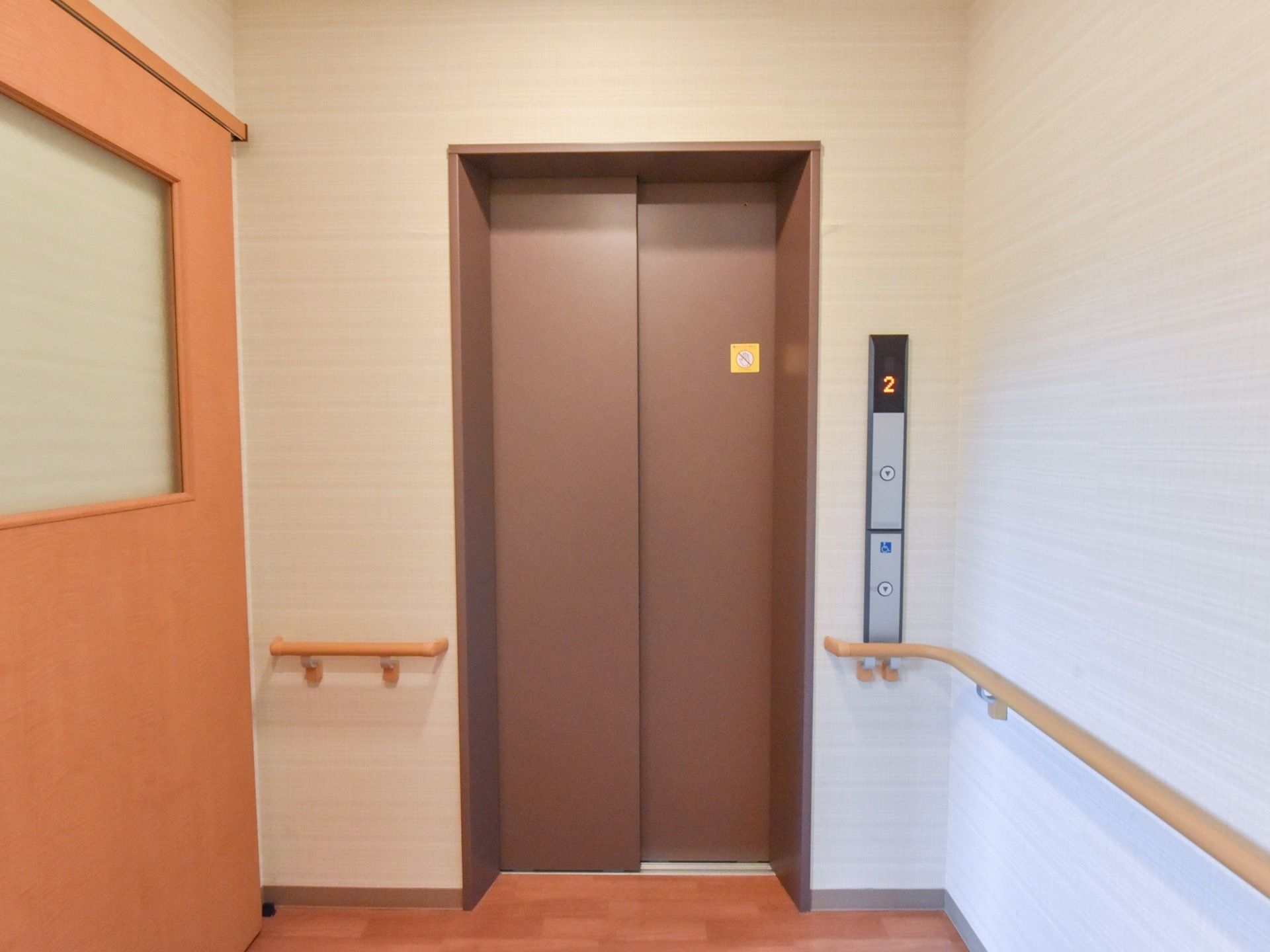通路の途中にはドアで仕切られた２階へのぼるエレベータがある。ボタンは低い位置に配置されており、手すりもある。