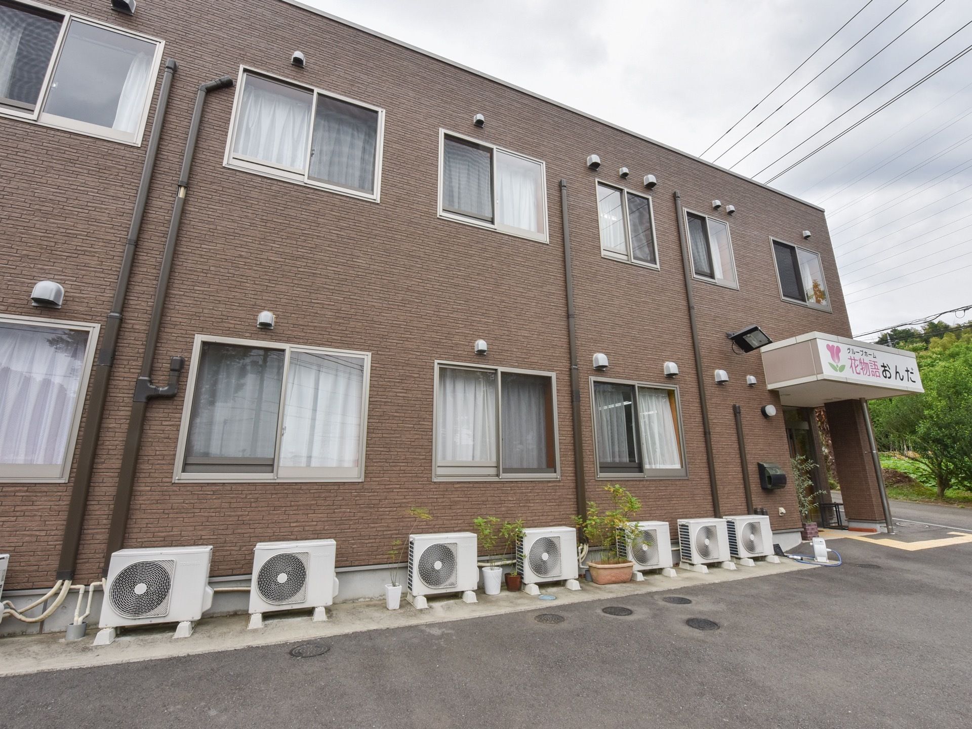 周辺は自然豊かな環境である。焦げ茶の外観の側面には間隔を置いて窓が並び、１階部分の地面の上に複数台の室外機を備えている。