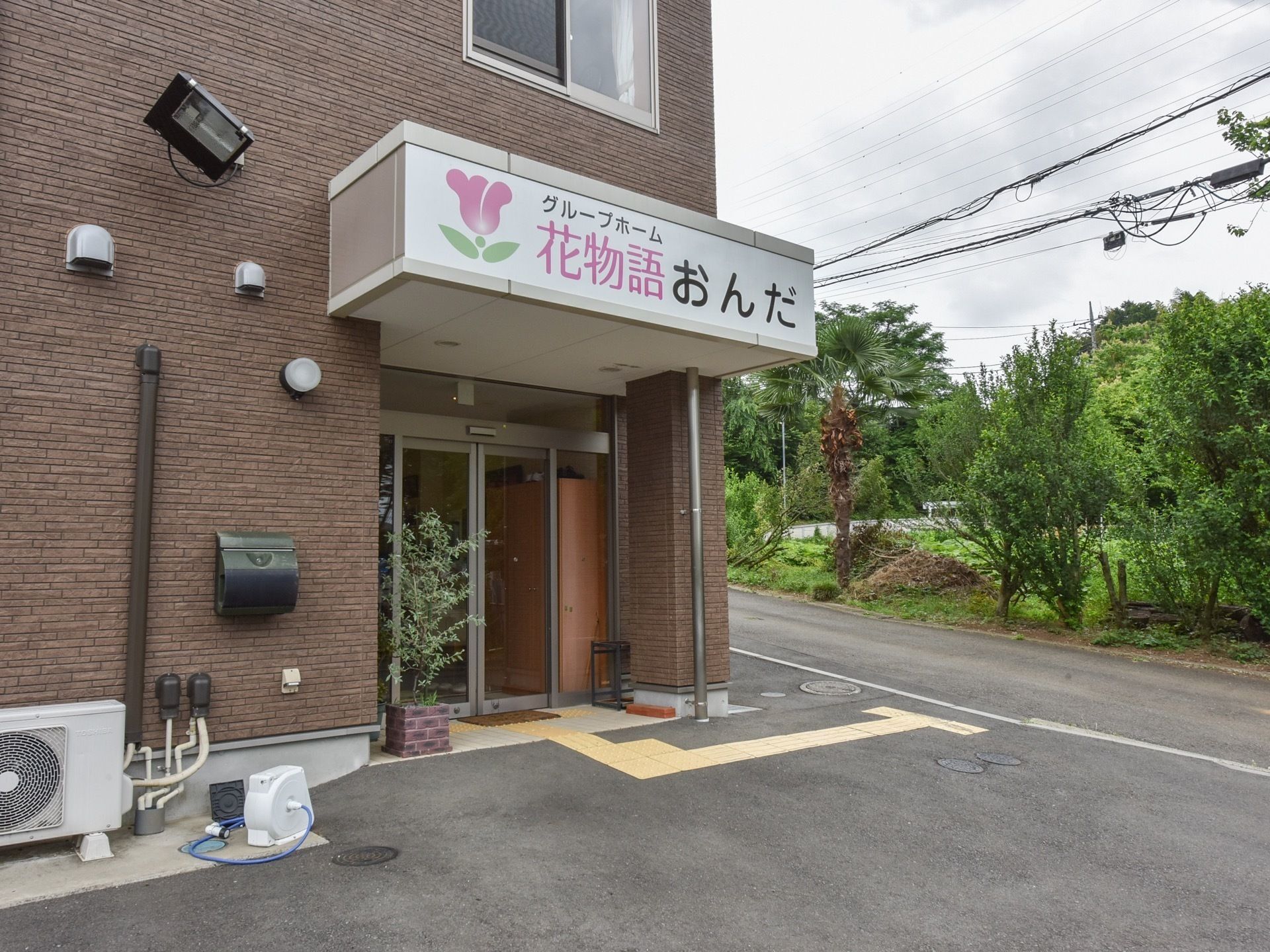 玄関の自動ドアの前に設けられた屋根には、施設名が大きな文字で記されている。足元には点字ブロックが備わっている。