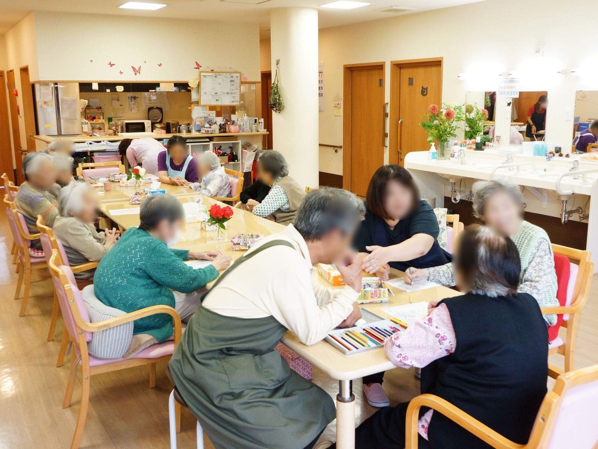 入居者様方が広々とした一室に集まり、塗り絵のレクリエーションを楽しんでいる。施設のスタッフが寄り添い、様子を優しく見守っている。