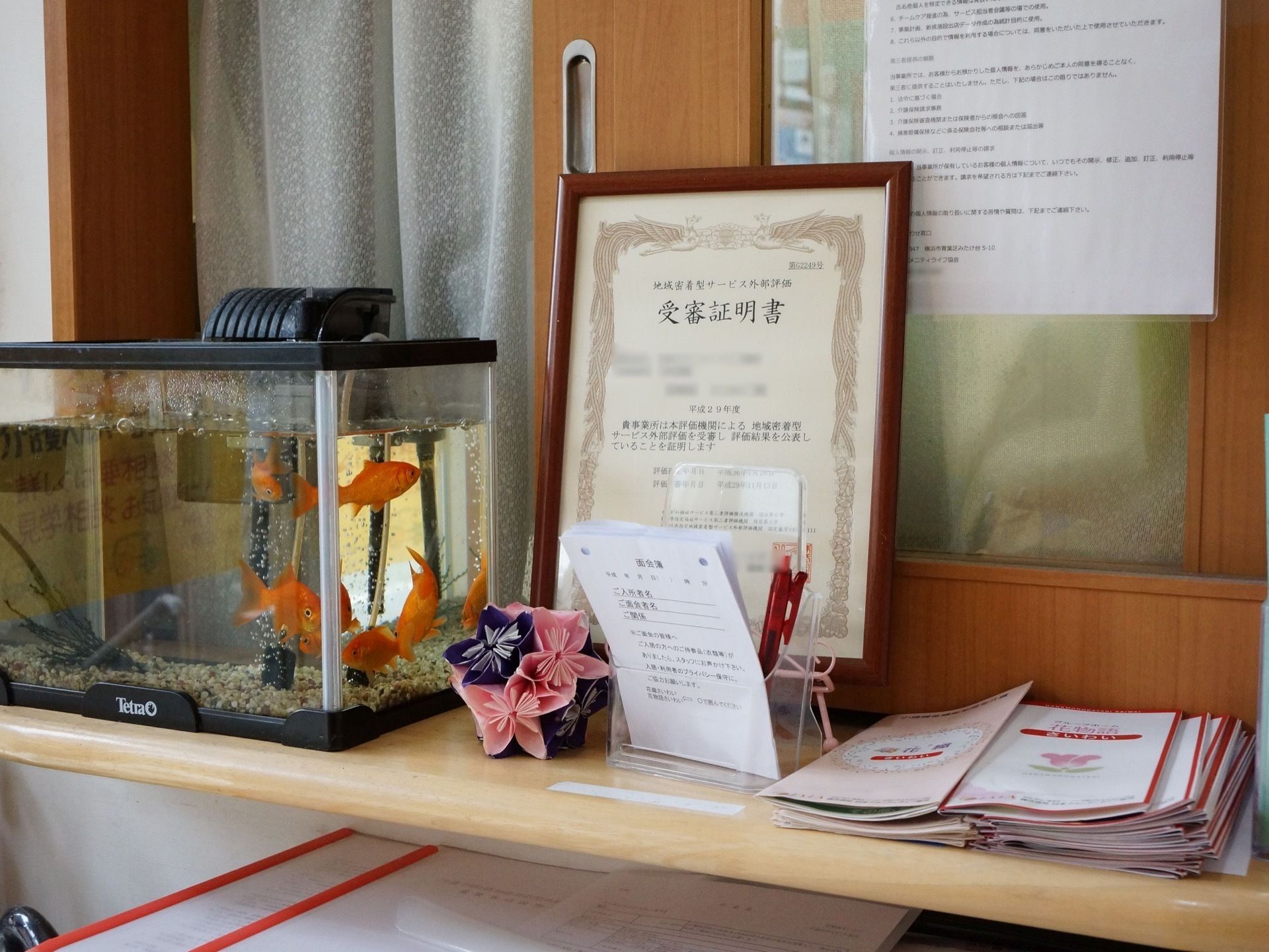 地域密着型サービス外部研修の受審証明書。金魚が気持ちよさそうに泳いでいる。面会のための記入用紙、施設のパンフレットなどが置いてある。