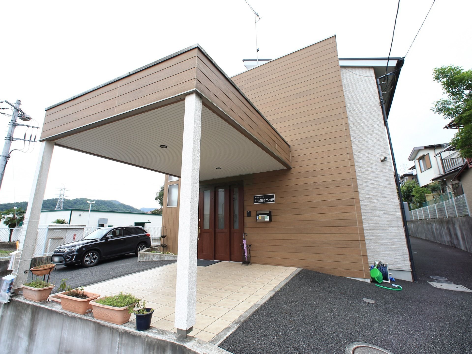 茶色の板張り風の外壁が落ち着いた佇まいの、木造２階建ての建物。ポーチの両脇が緩い傾斜で、雨天の場合も足元が悪くならないように配慮されている。