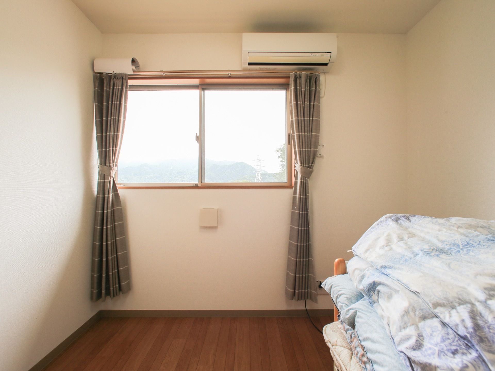 少し高い位置にある窓には、裾の長い厚手のカーテンが取り付けられている。手前には布団が畳んで積まれている。