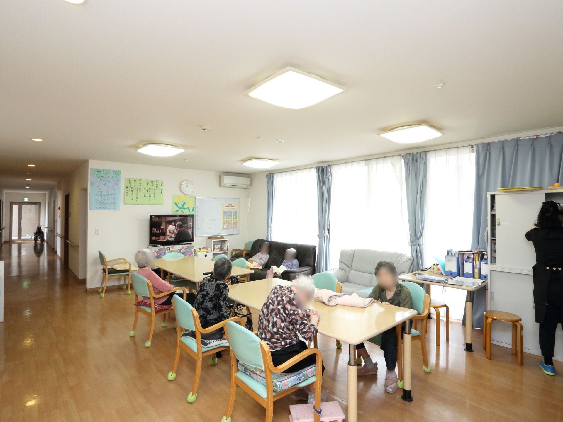 大きなガラス戸から自然光が差し込む広い空間には、ダイニングテーブルと椅子が配置され、入居者様が着席して壁際のテレビに視線を向けている。