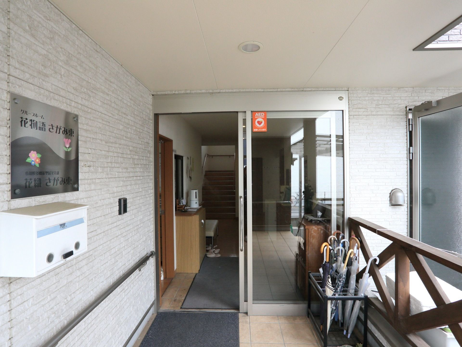 玄関入口前には施設名が書かれたシルバーの看板が壁に貼られている。下にはポストがある。インターフォン、手すりもある。