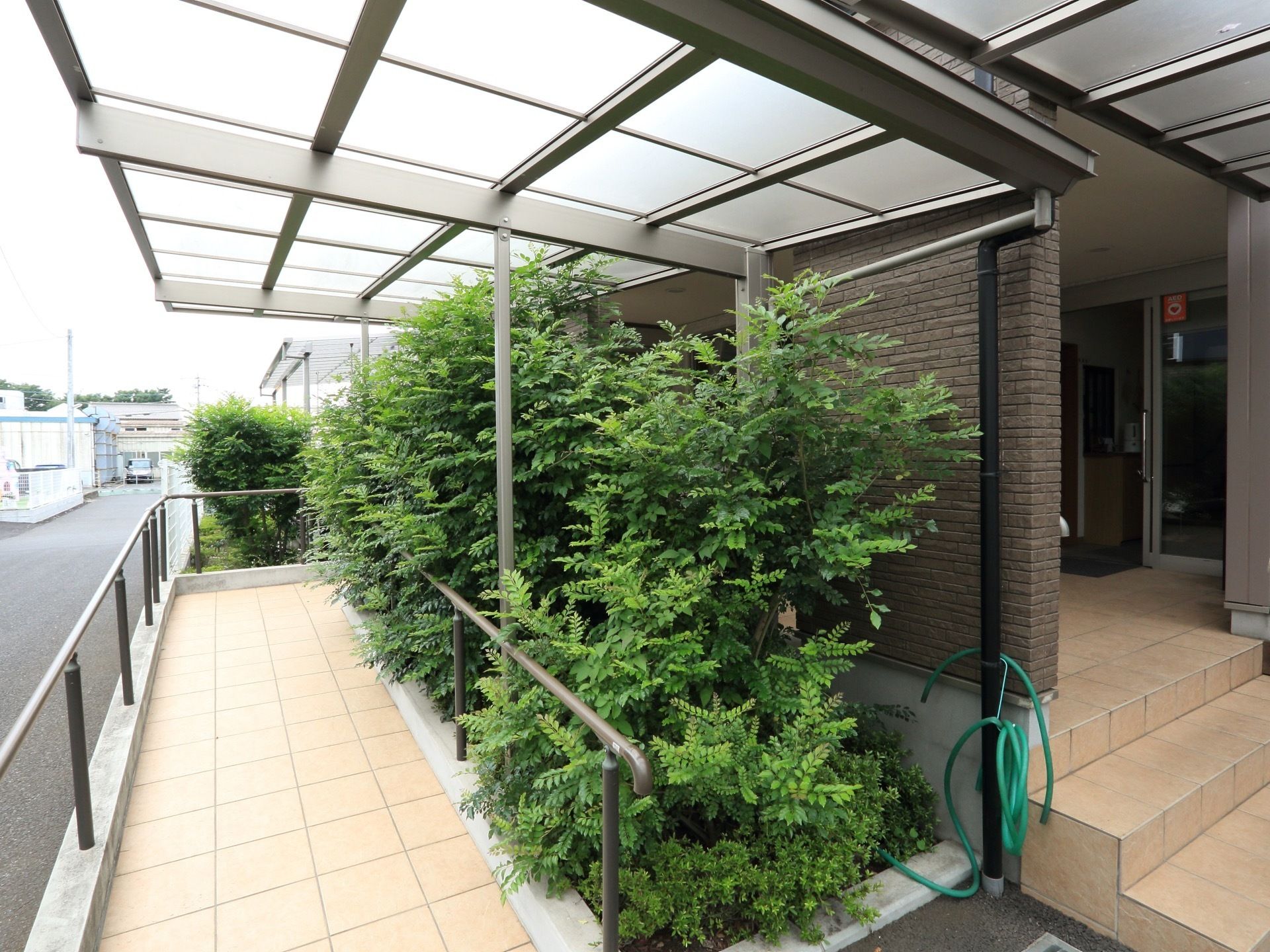 施設前には大きな屋根が取り付けられている。その下はスロープになっている。両側には手すりがある。植え込みには緑が植えられている。
