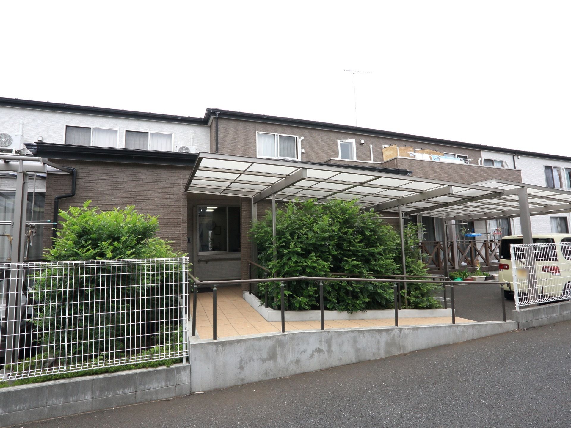 施設は２階建てである。両側に手すりが付いたスロープの上には屋根がある。横には車が１台停められている。