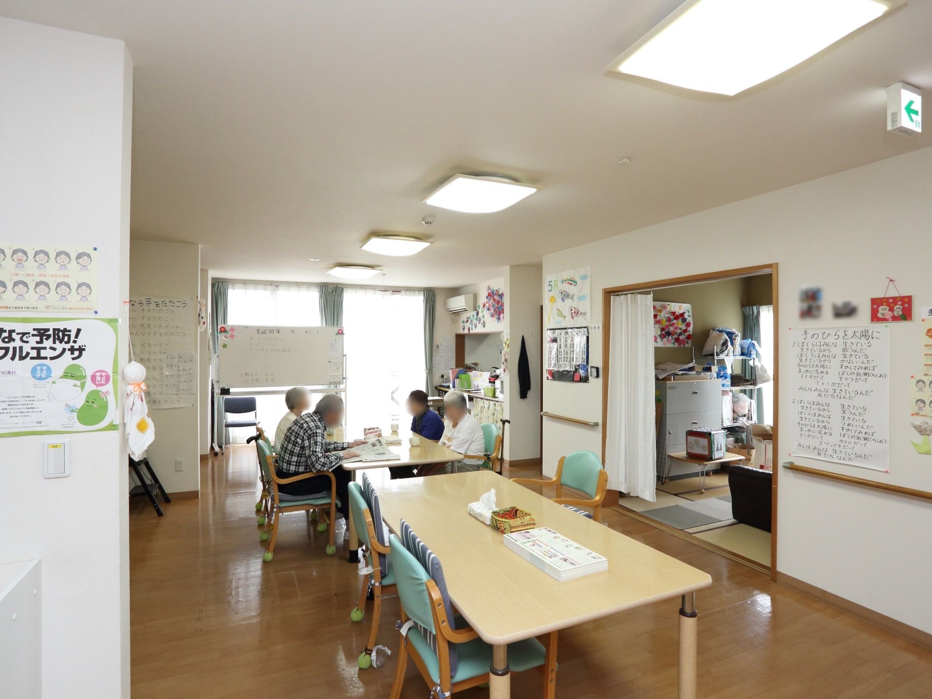 食堂には２台のテーブルがあり、１台のテーブルには入居者様が４人集まって座っている。壁に幸せなら手をたたこうの歌詞が書かれた紙が貼られている。