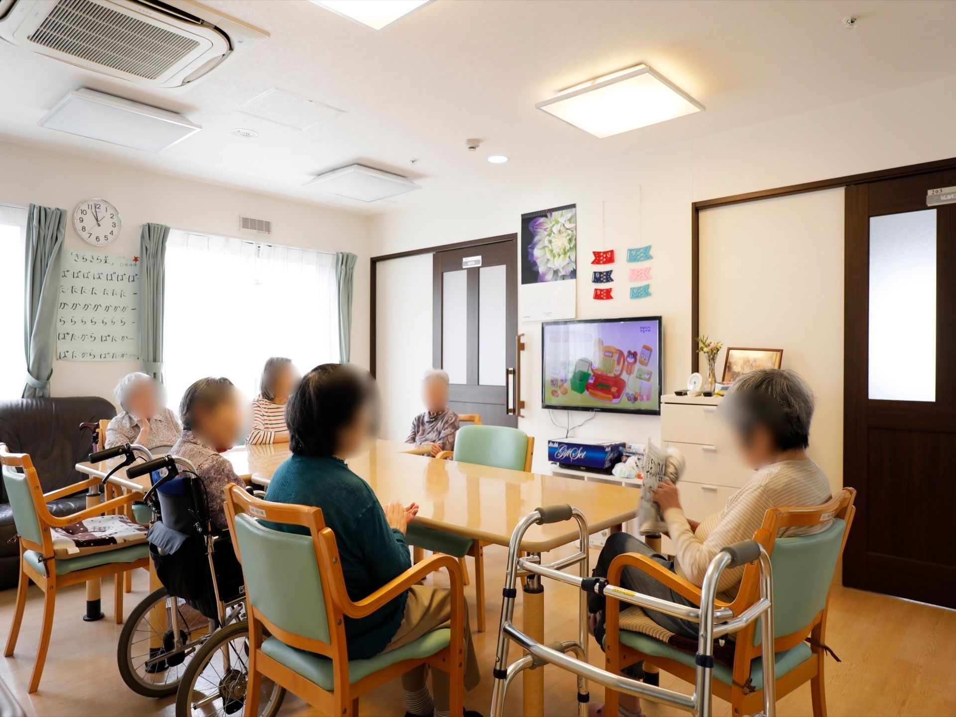 ダイニングテーブルを囲んで車椅子をご利用の方や椅子に腰を掛けた入居者様が、壁掛けの薄型テレビを視聴している。