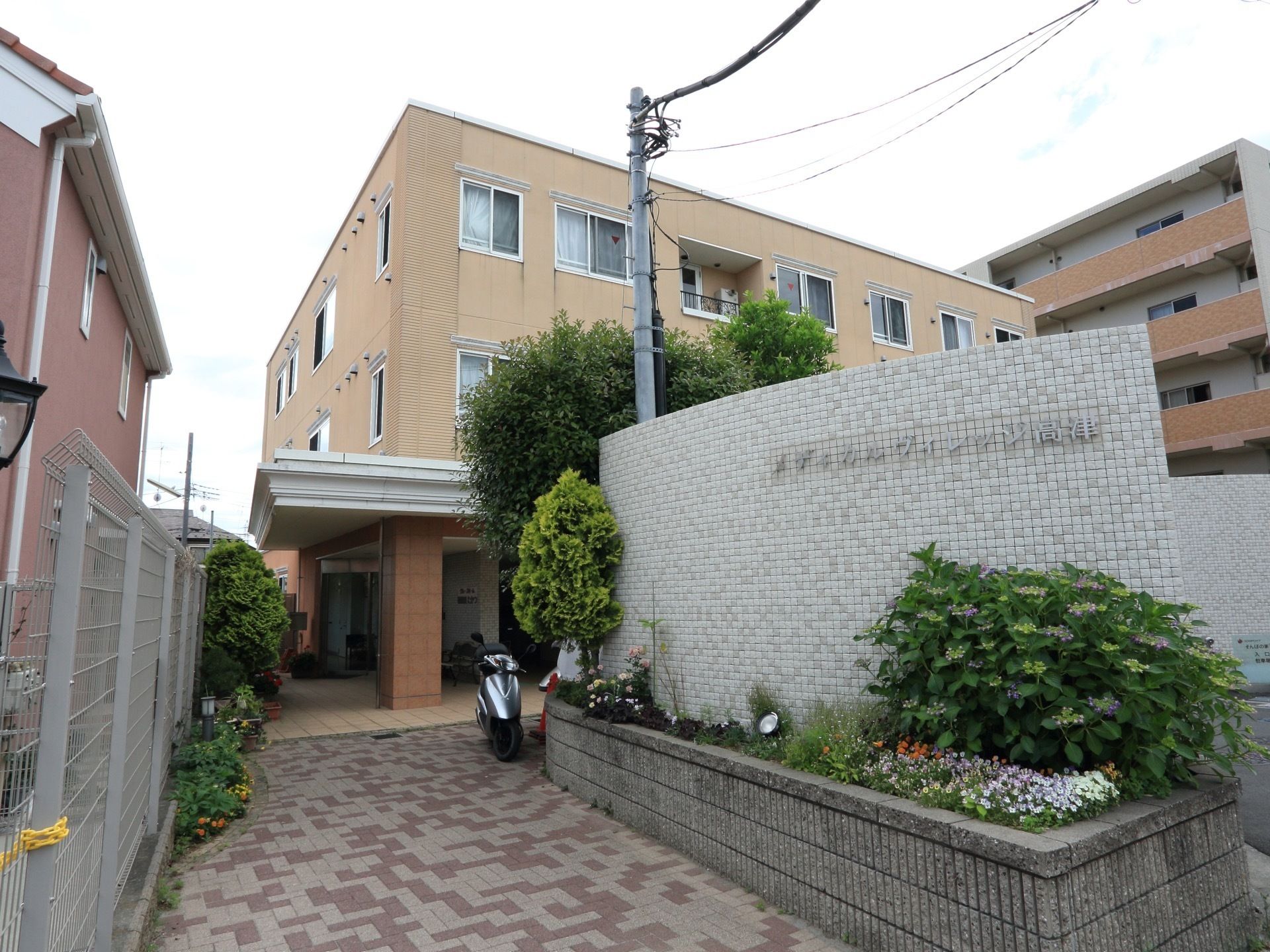 外観はベージュ色の壁。3階建ての建物。玄関への通路は広く取られている。花壇にはさまざまな植物が植えられている。