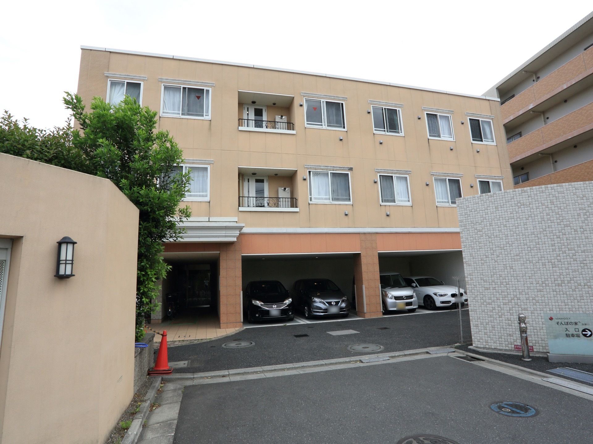 オレンジ色の壁が特徴のマンションタイプの建物。車が入りやすい広めの門の脇には自然を楽しめる緑が植えられている。