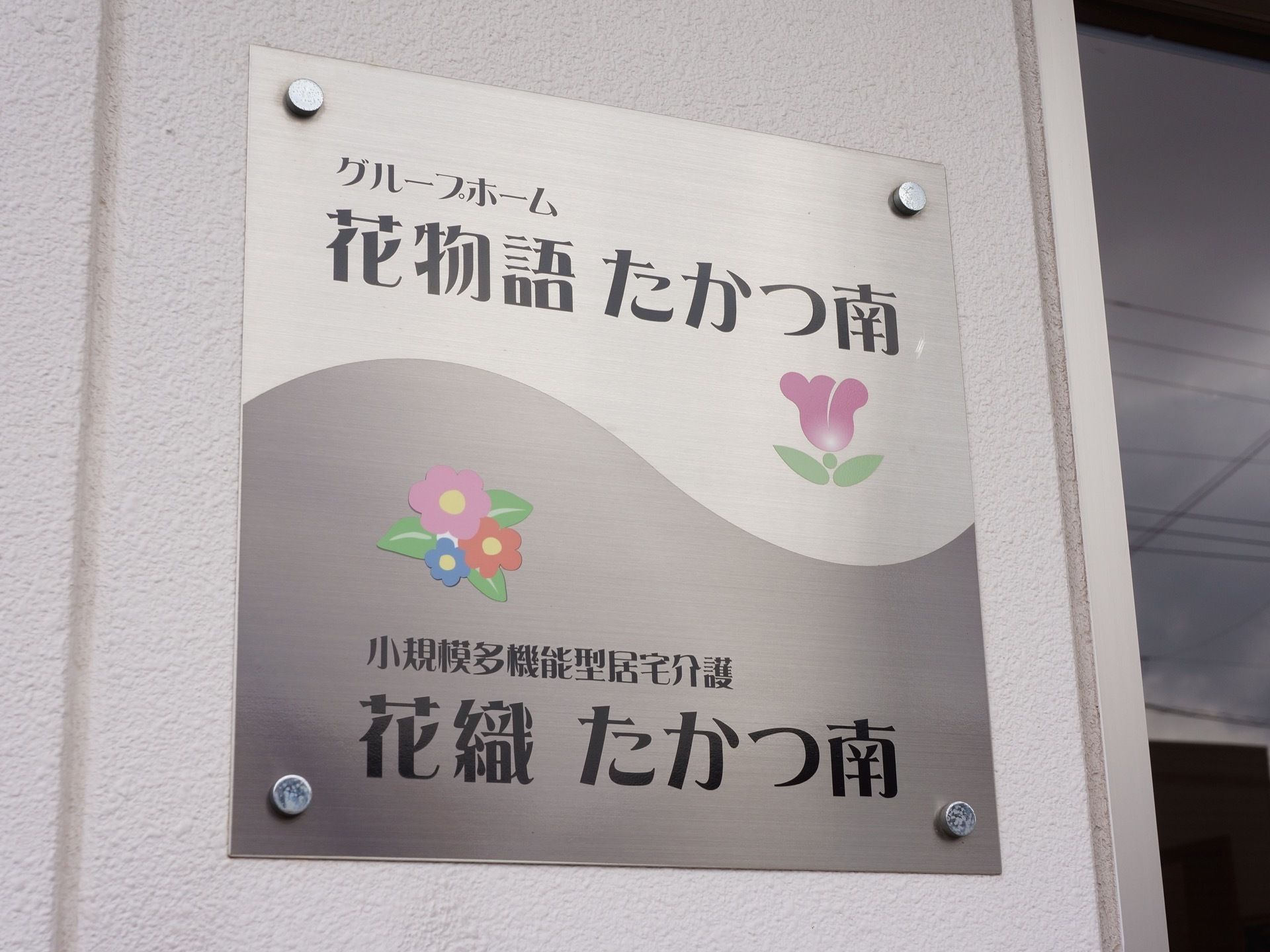 外壁に金属製の看板を設置している。当施設の名称のほかに、同一建物内に併設しているサービス名と合わせて表記している。