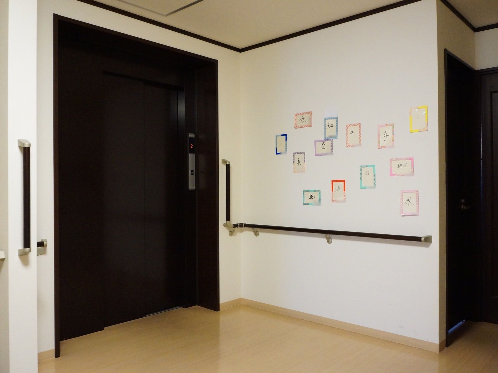 手すりが設置された白い壁とダークブラウンのエレベーターの扉がある。壁には入居者様の作成したカードが飾られている。