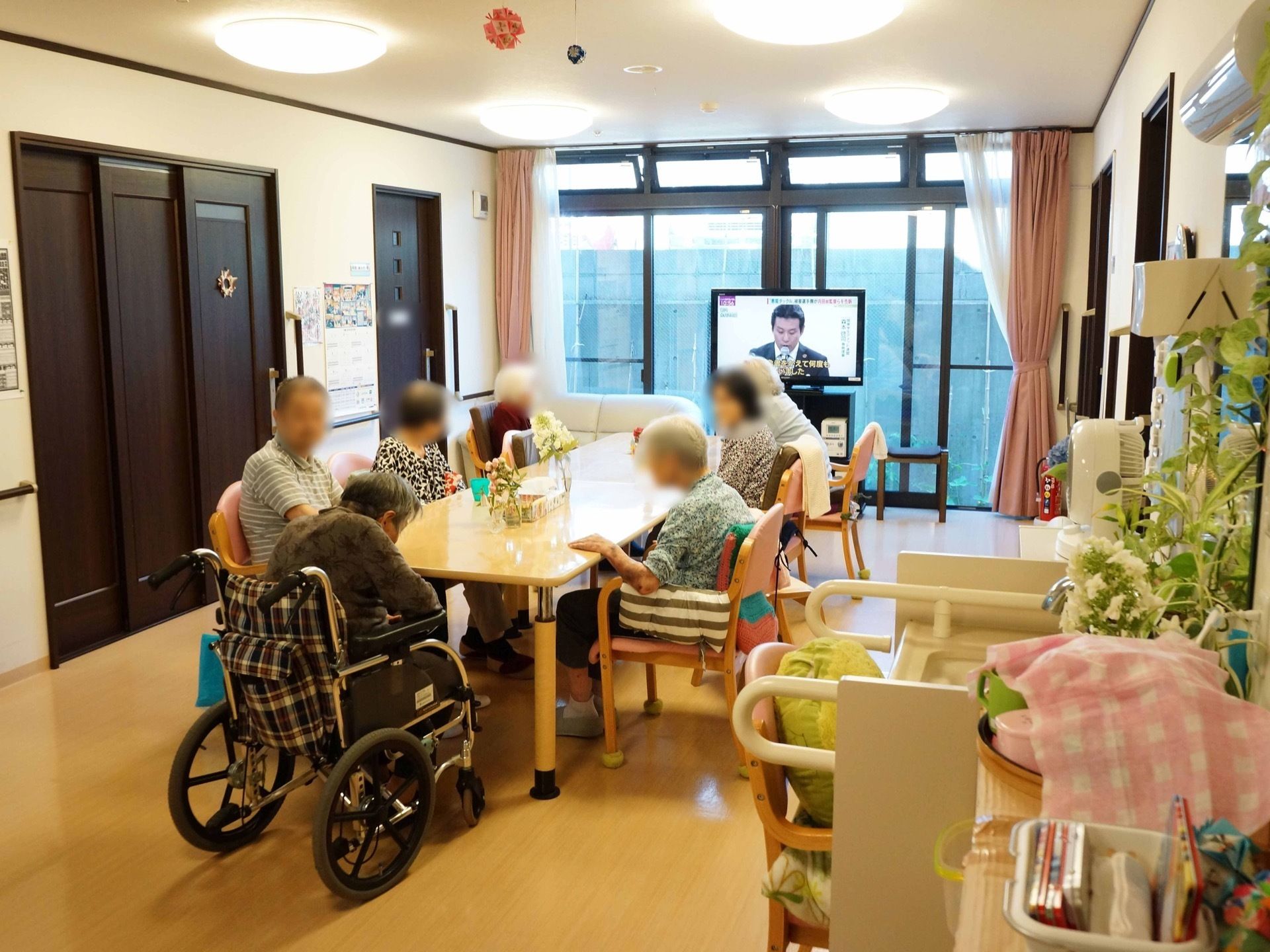 広々した長方形のテーブルが設置されている。入居者様が椅子に座って思い思いの時間を過ごしている。窓側にはテレビがある。