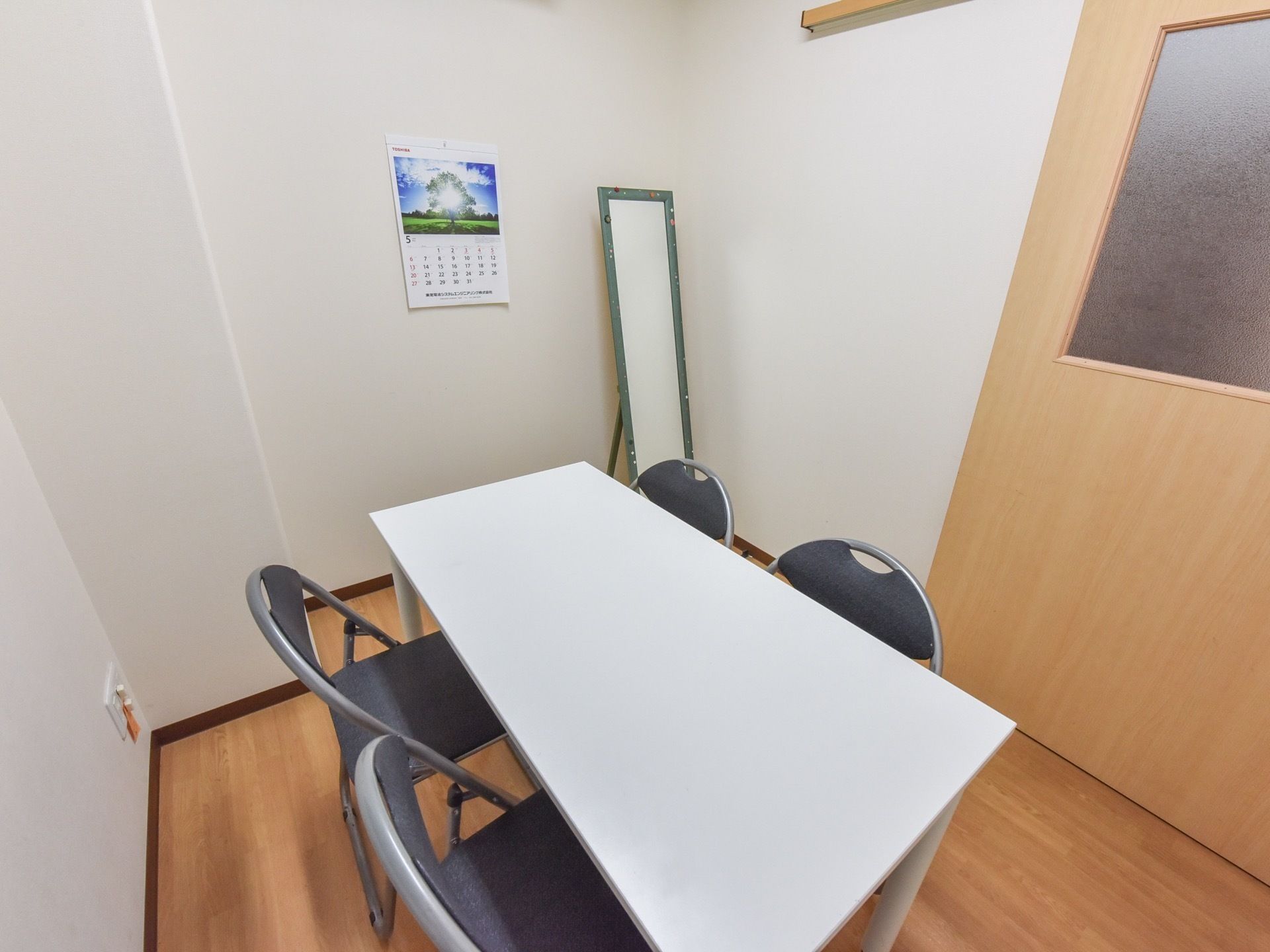 室内にはテーブルとパイプ椅子が配置され、入口の横にはスタンドミラーが置いてある。壁にはカレンダーが貼られている。