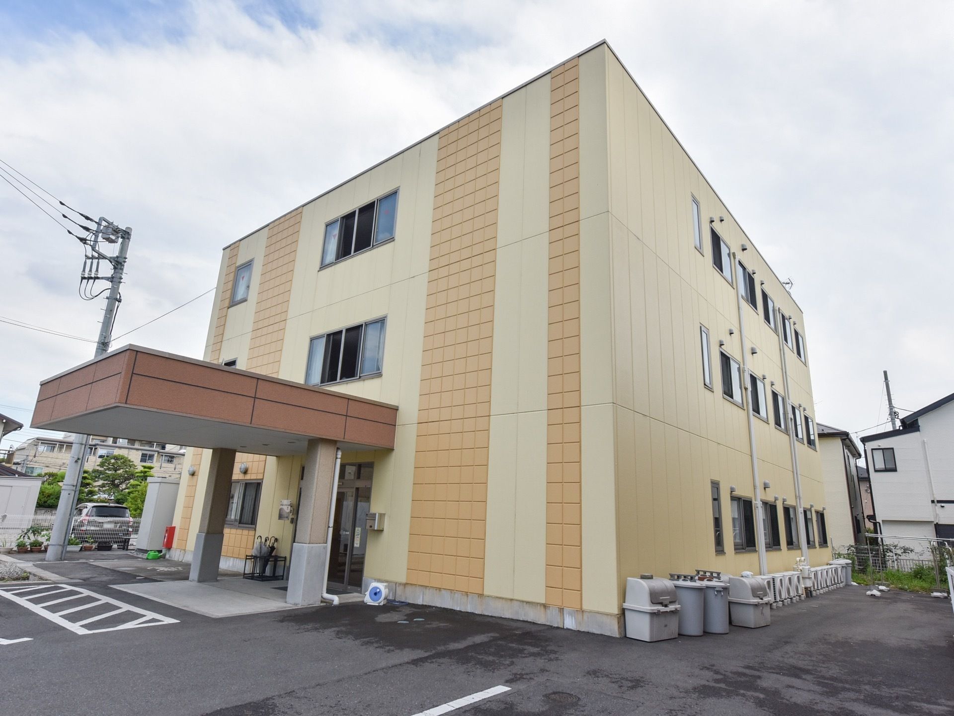 濃淡２色のベージュ色の外壁。建物は３階建で四角い形をしている。周囲はアスファルトの駐車場となっている。