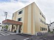 濃淡２色のベージュ色の外壁。建物は３階建で四角い形をしている。周囲はアスファルトの駐車場となっている。
