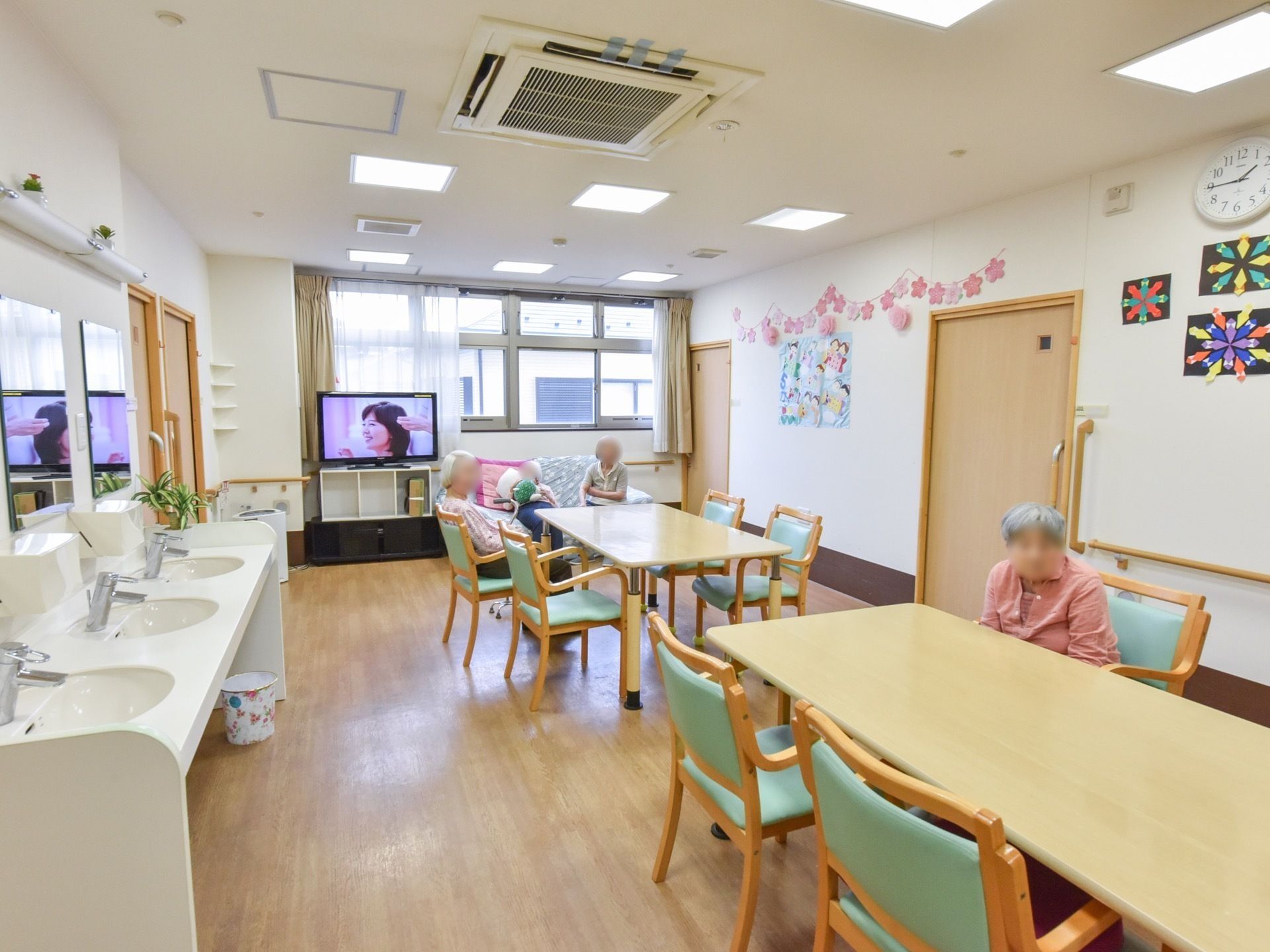 左手に洗面台が３つある長方形のリビングルームでそれぞれに寛ぐ入居者