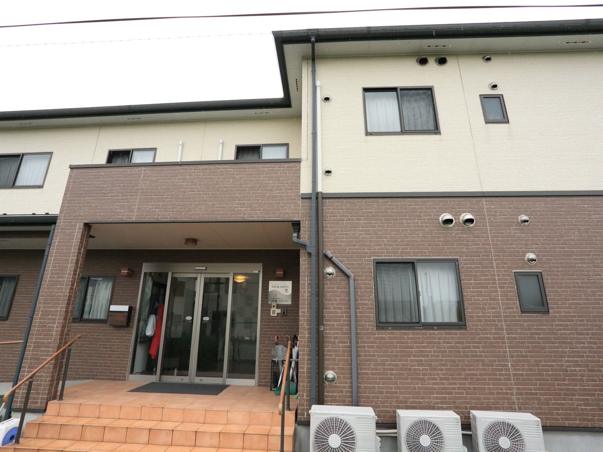 １階部分がブラウン、２階部分がアイボリーの優しい色合いの外壁が特徴の建物。入口には屋根が設置されている。