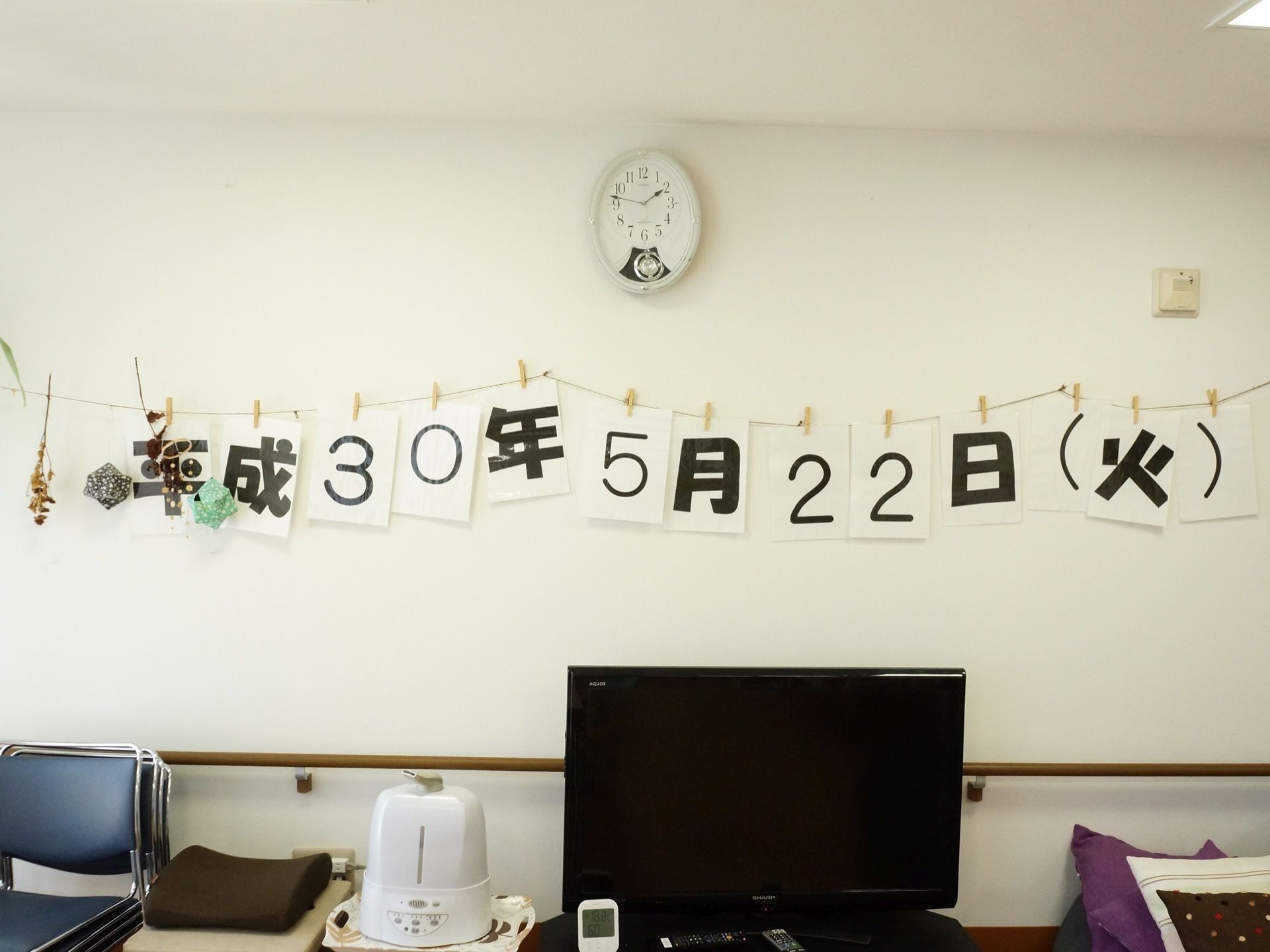 共有スペースの白壁に１枚の用紙に１文字ずつ書いて横に並べ、本日の日付と曜日を表示している。その上に時計を掛け、下にテレビを置いている。