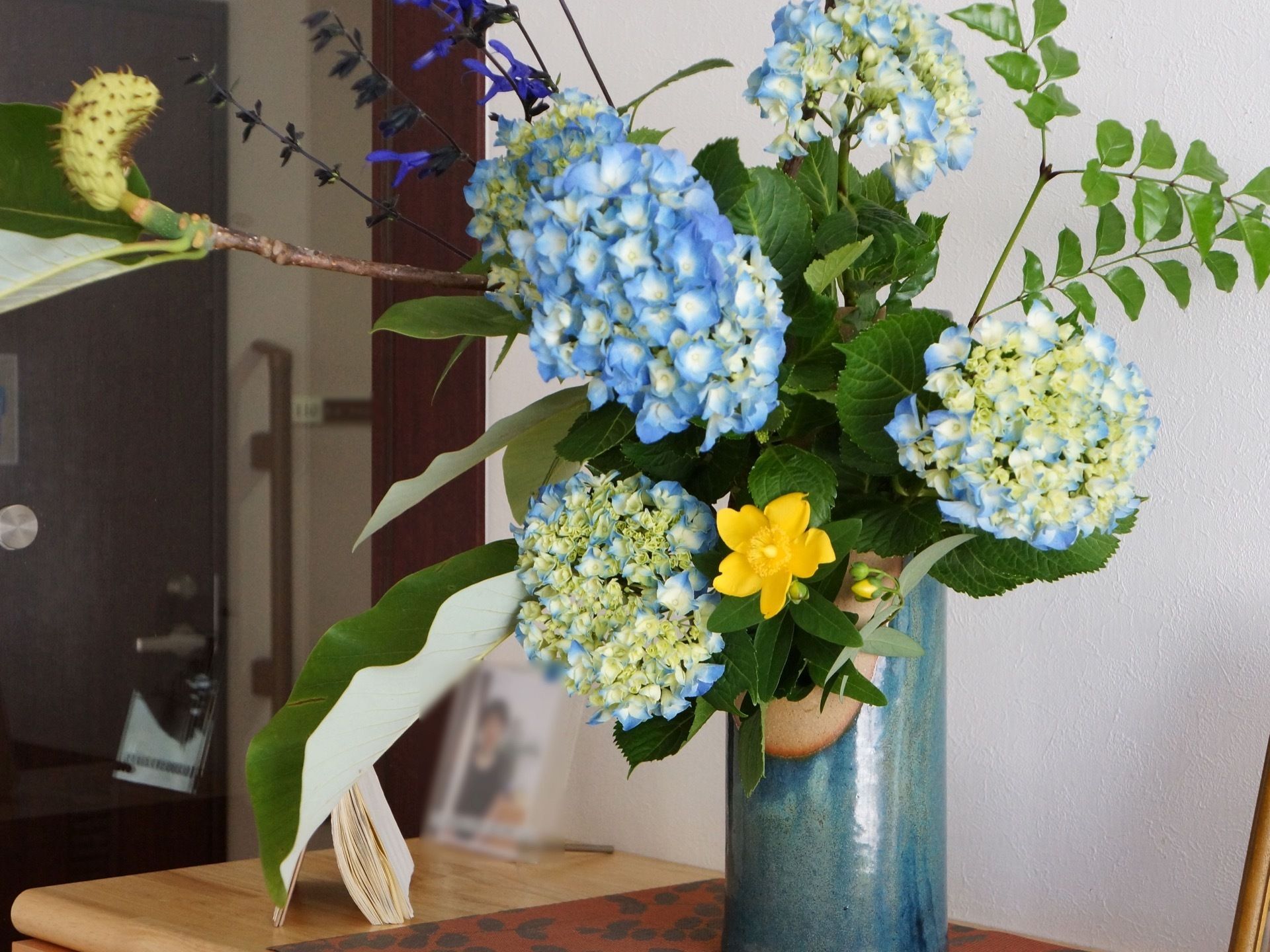 壁際の棚の上に陶器の花瓶に飾られた紫陽花と黄色い花がある。本格的な生け花の雰囲気があり、見る人を和ませる。