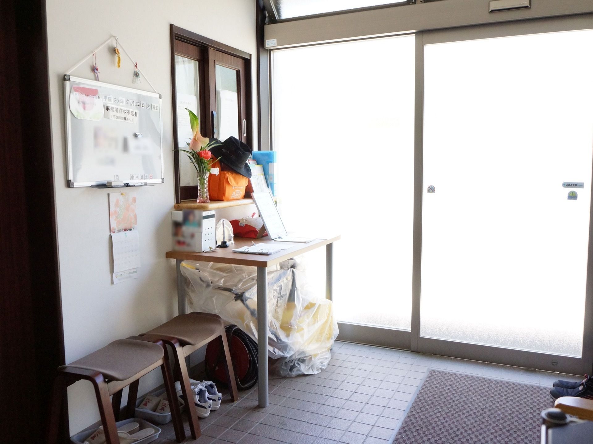 ガラス張りで自然光が差し込む明るい自動ドアの玄関がある。壁際には椅子とお知らせ用の掲示板ボードと机が設置されている。